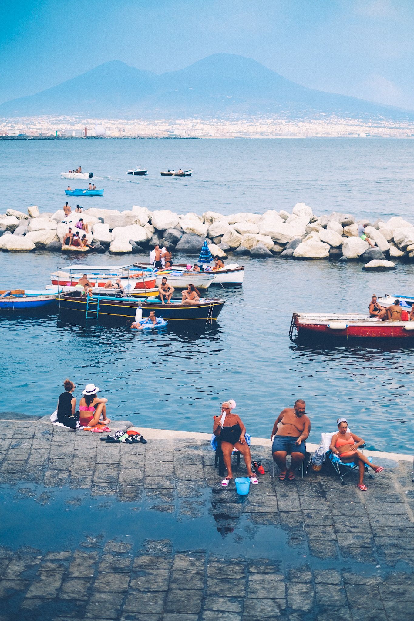 People sit on the pier of Napoli, boats in the water and Mount Vesuvius in the background, faint.
