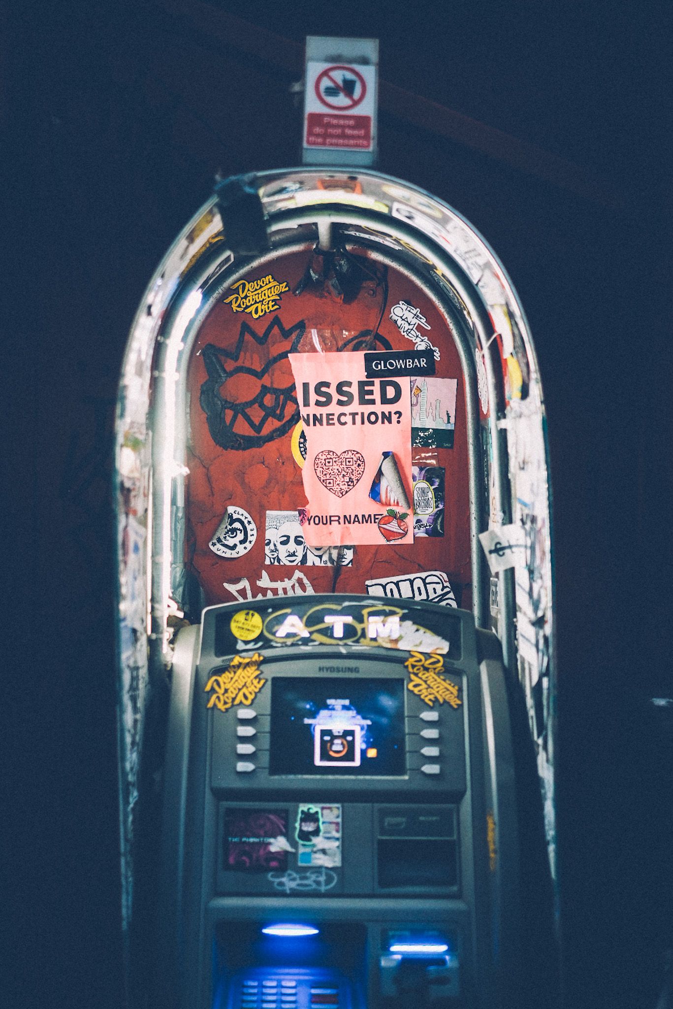 An outdoor ATM machine with stickers and graffiti sits in the dark, with a torn “missed connection” paper hanging in the center.