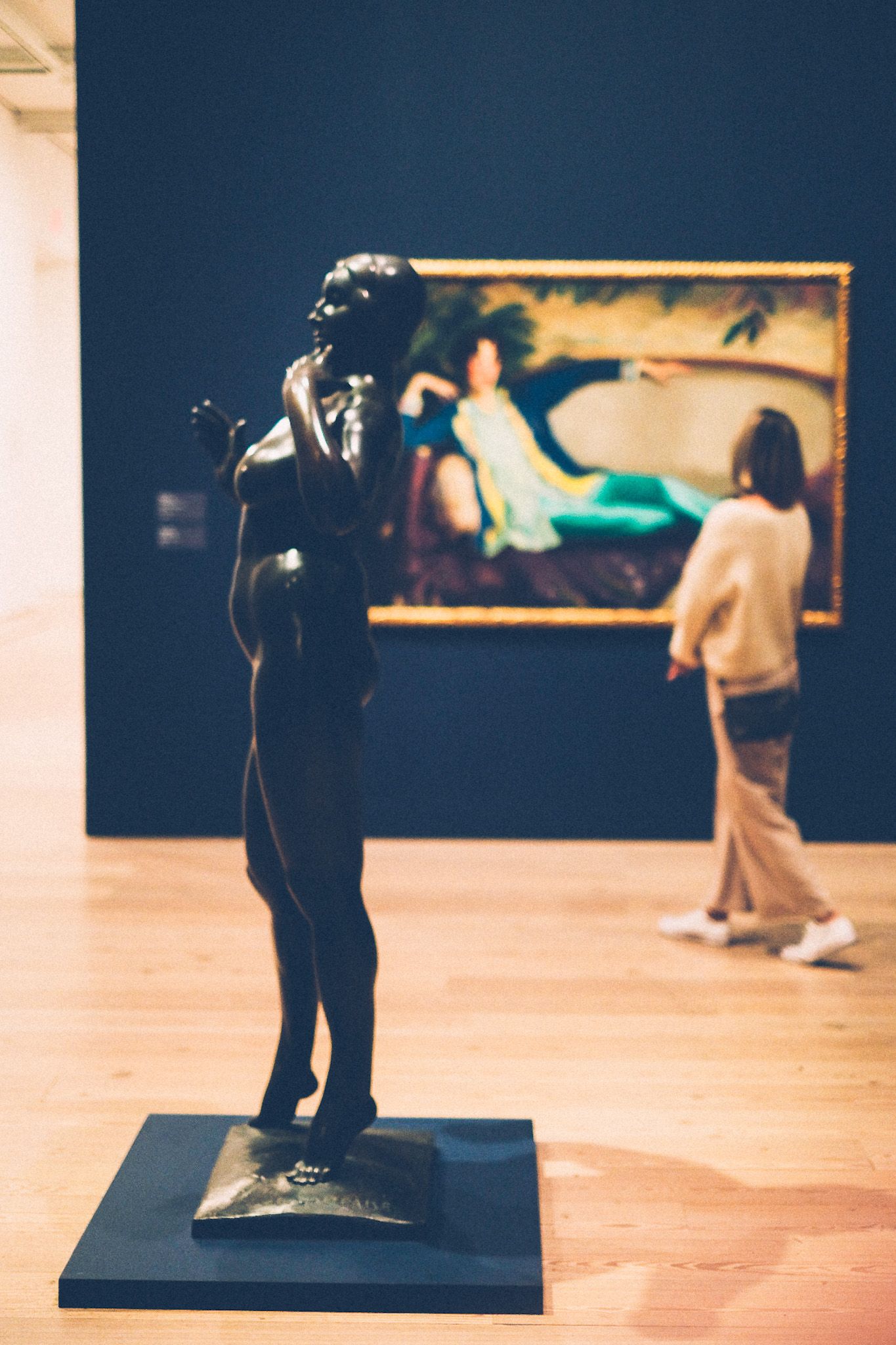 In a museum, a brass statue of a nude woman stands on the left side of the photo, showing her side profile, while in the background a person looks at a large painting of a woman lounging. The gallery wall is painted dark navy.