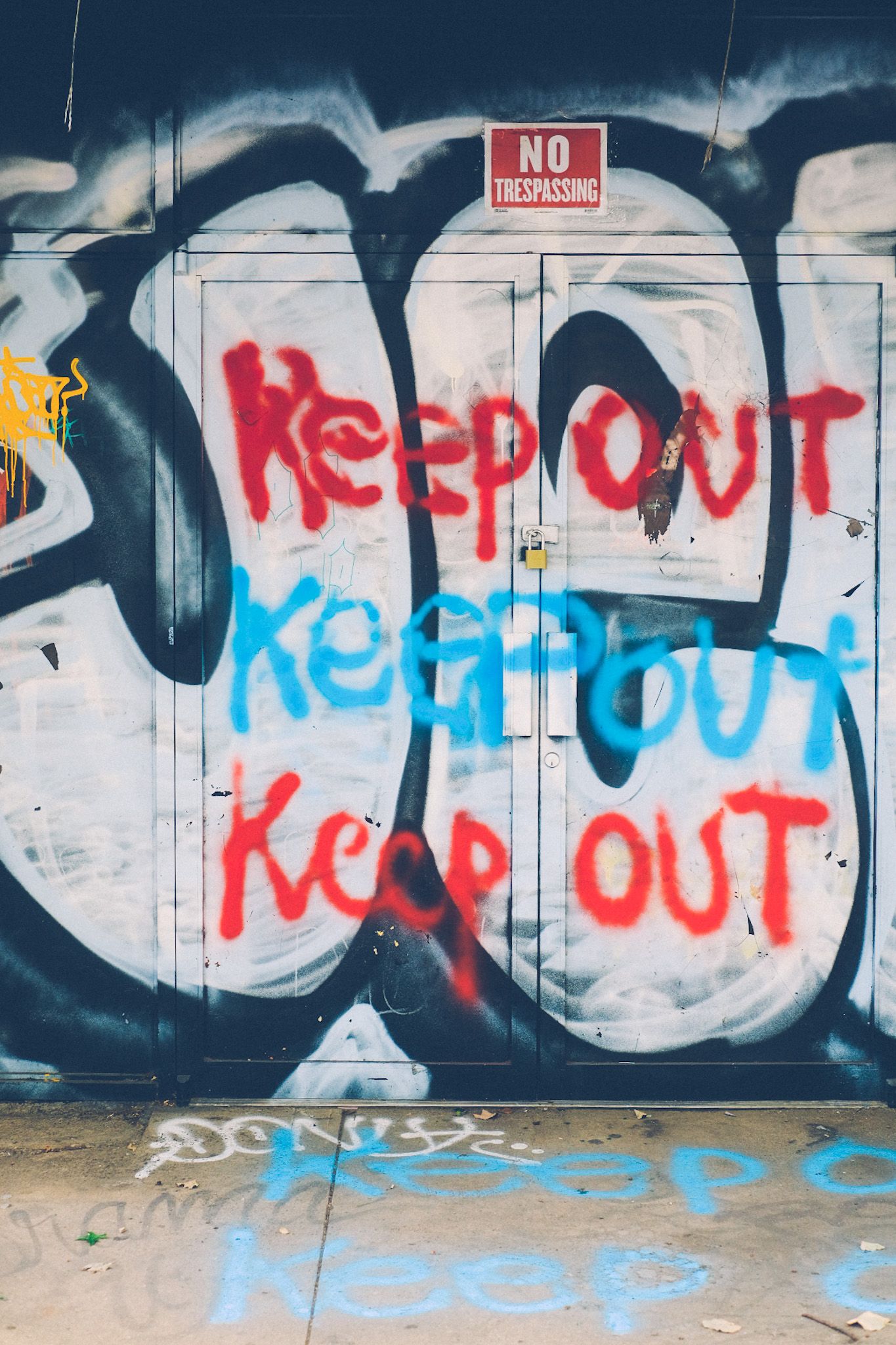 Graffiti across the front doors of a building repeats the words “KEEP OUT” three times in alternating blue and red, also repeated across the sidewalk.