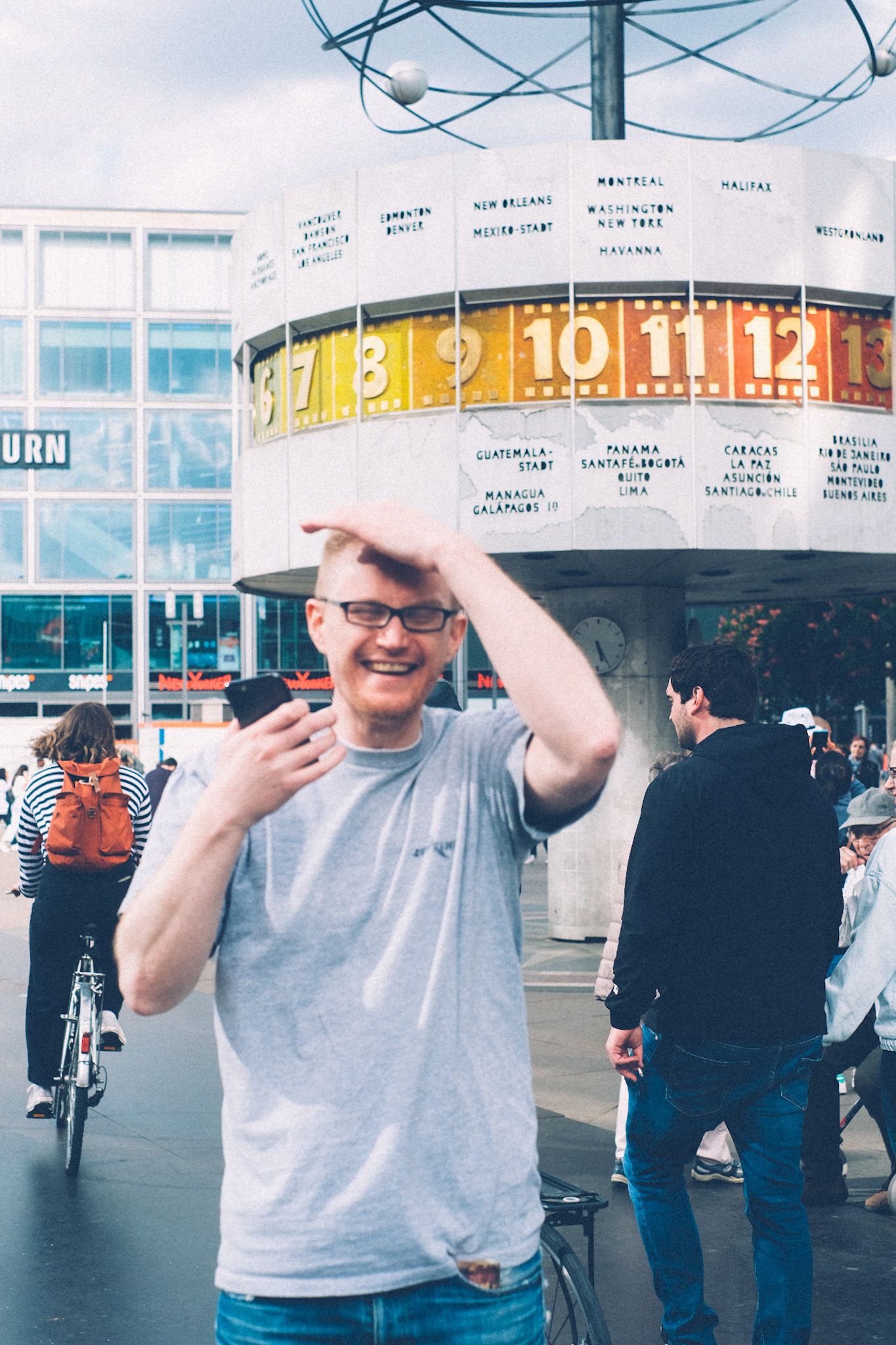 A man stands in front of the world clock installation in Berlin, smiling in front of crowds, grabbing his head with his hands.