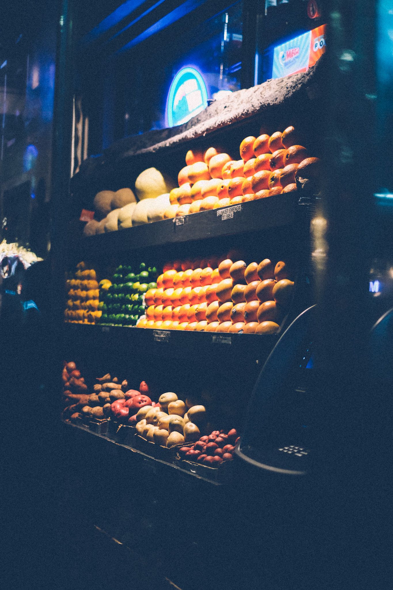 Stacks of citrus fruit sit on the shelves outside of a bodega, illuminated by one overhead light. It is evening.