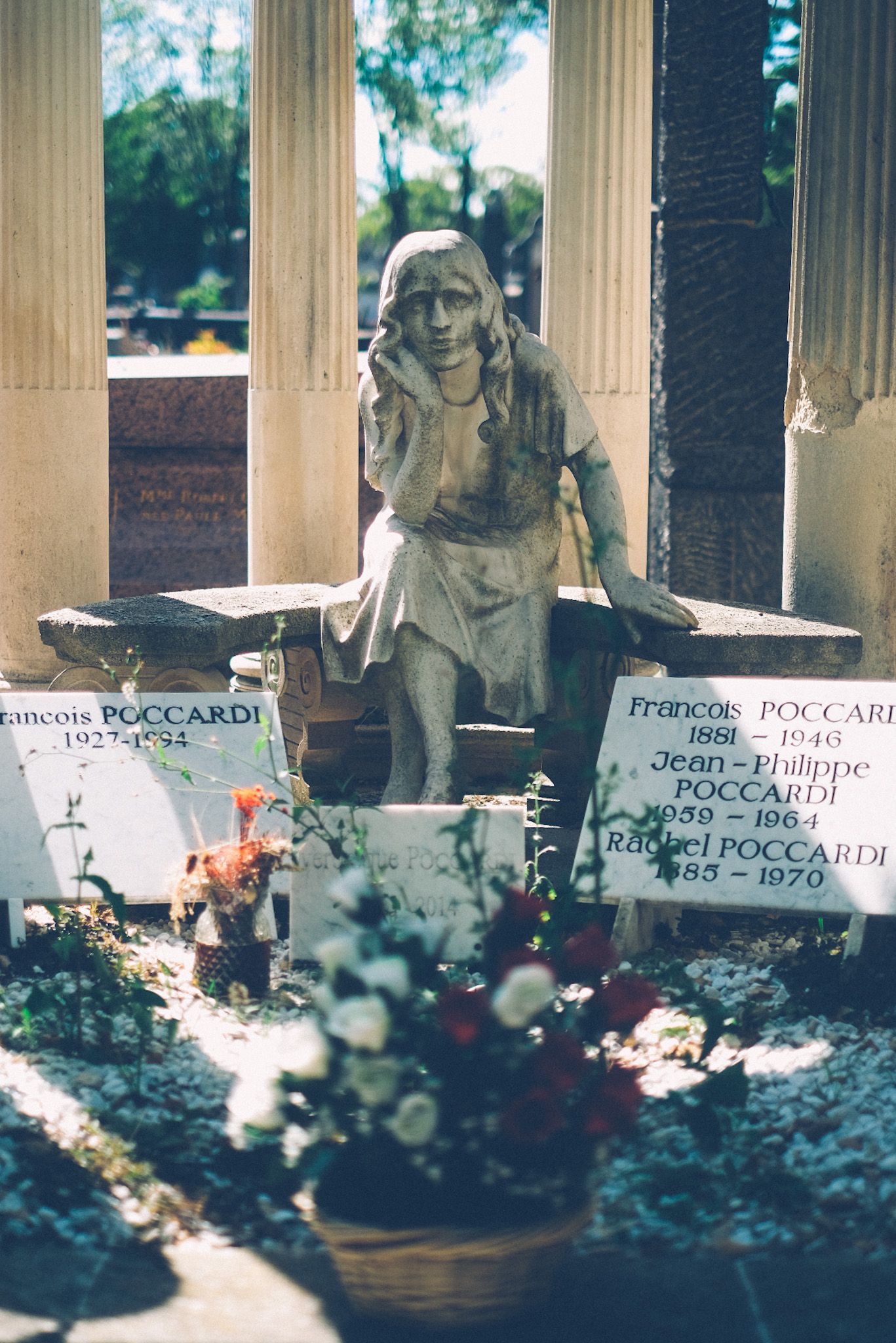 A statue atop a grave in Pére Lachaise cemetery holds its chin in its hand as if looking into the camera. Behind it are four columns, beside it are gravestones with French names, and in front is a bouquet of flowers slightly out of focus.