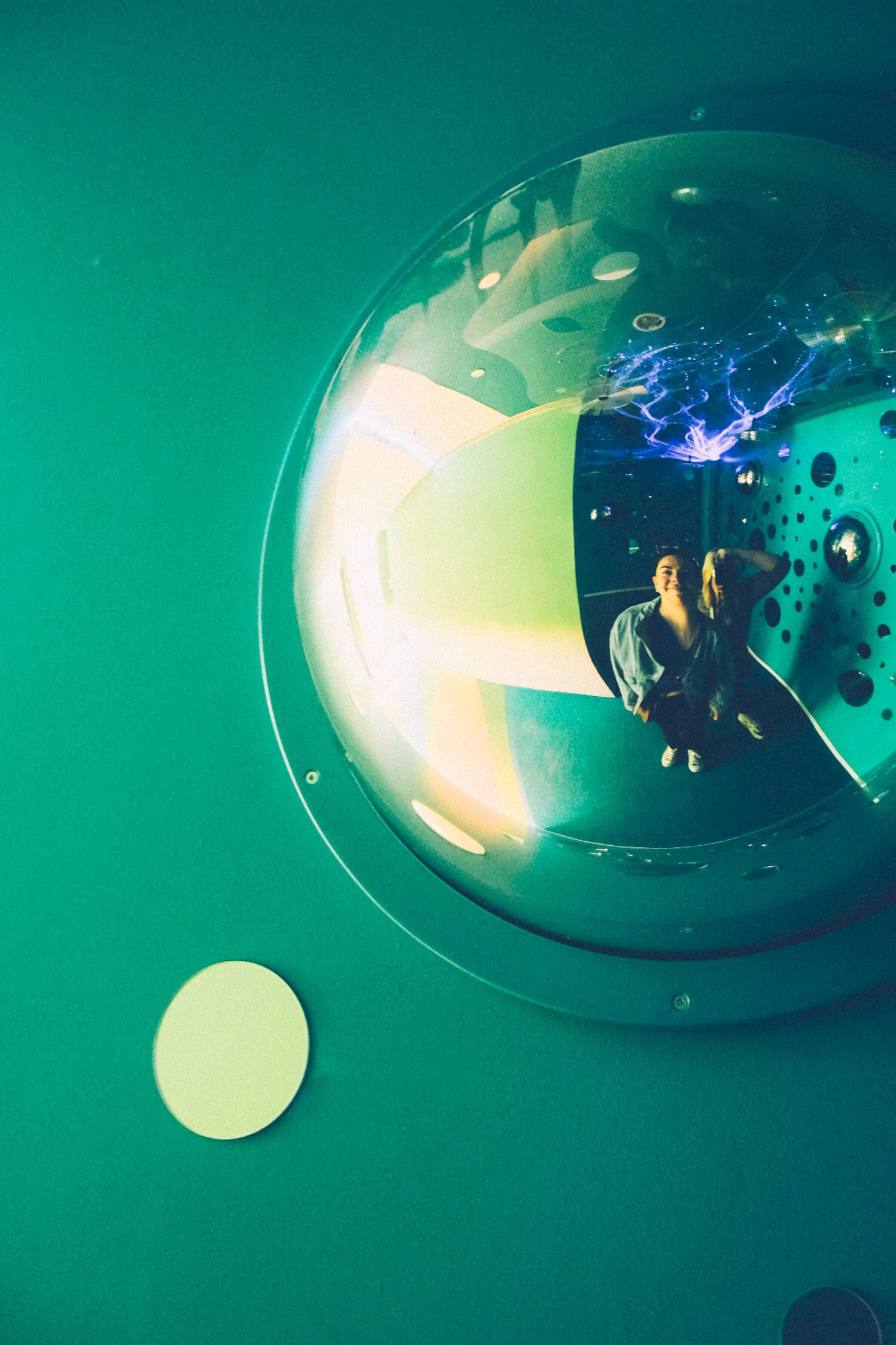 Two women are reflected in a chrome bubble jutting out from a green wall in the children’s section of a museum. Their images are warped in the sphere.