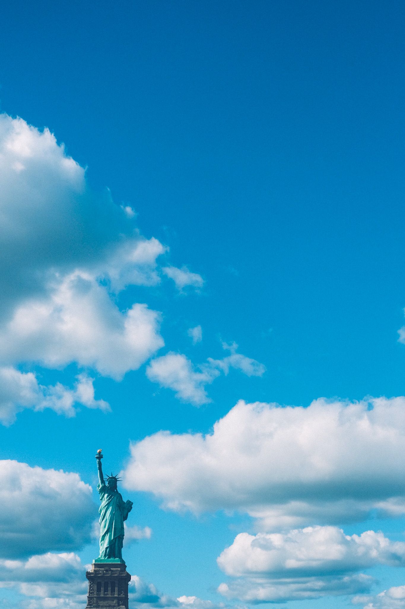 Amid a background of partly cloudy blue sky sits the Statue of Liberty, rising small from the bottom left corner.