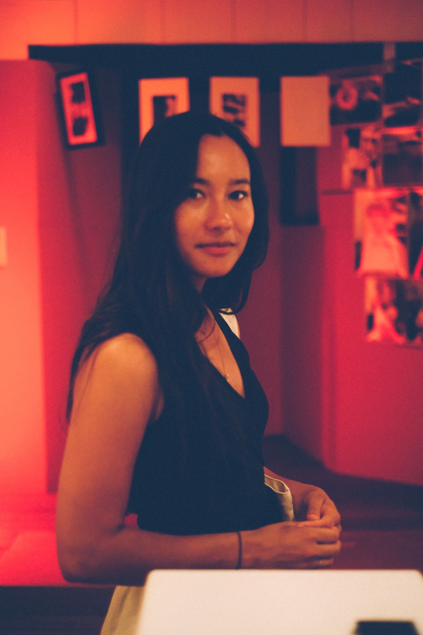 A woman with long, dark hair stands mid-frame in a room full of red, full of dark photographs, looking at the camera.