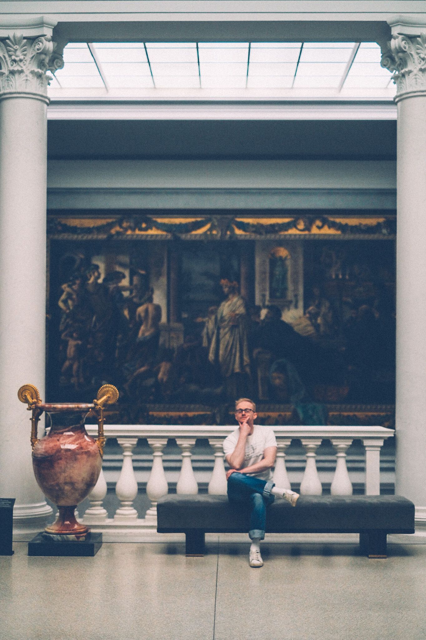 A man sits on a bench in a classic art museum, next to a large vase and in front of a mural, his hand on his chin.