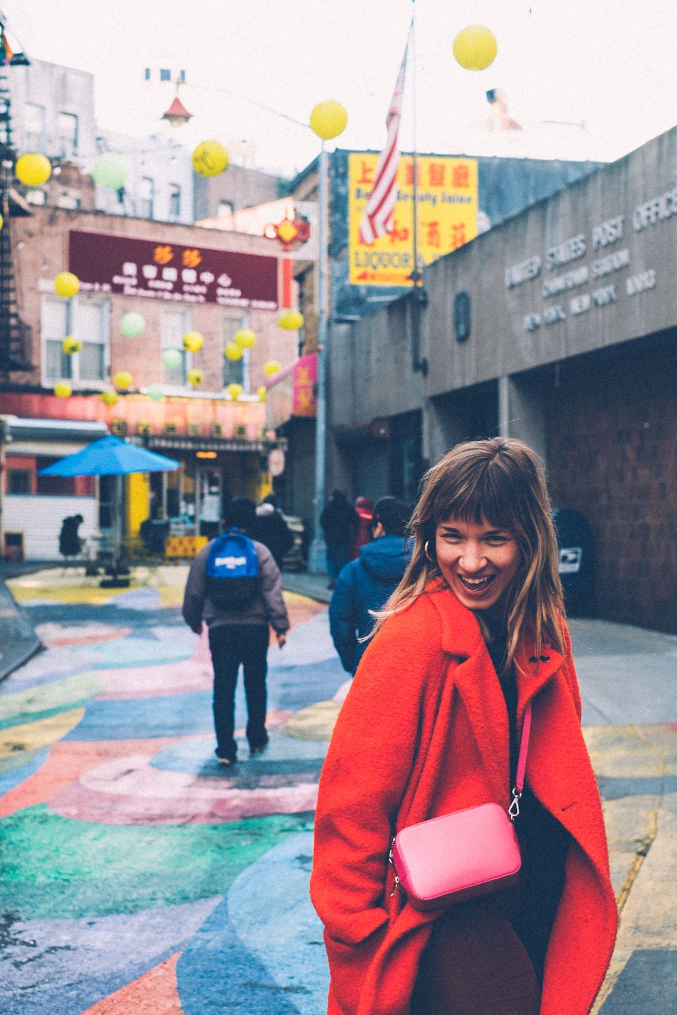 A woman in a red coat with a pink purse smiles, posing in front of a colorfully painted street in Chinatown. Yellow lanterns hang over the walkway and people mill about in the background.
