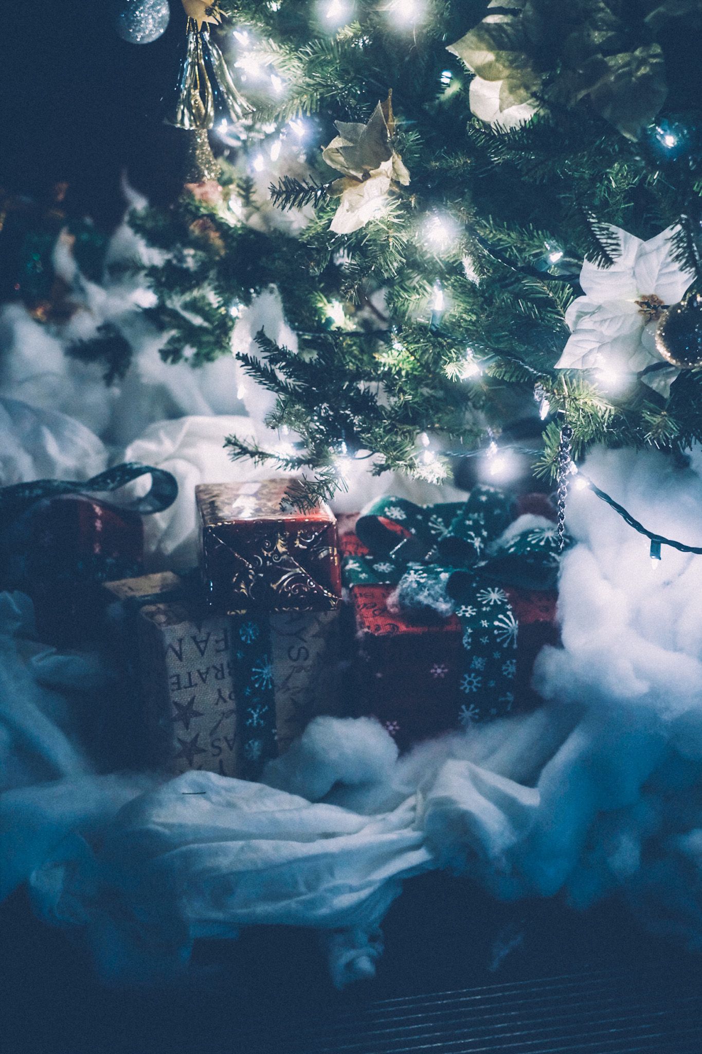 A pile of Christmas presents sits under a decorated tree with soft white lights, all atop fake snow.