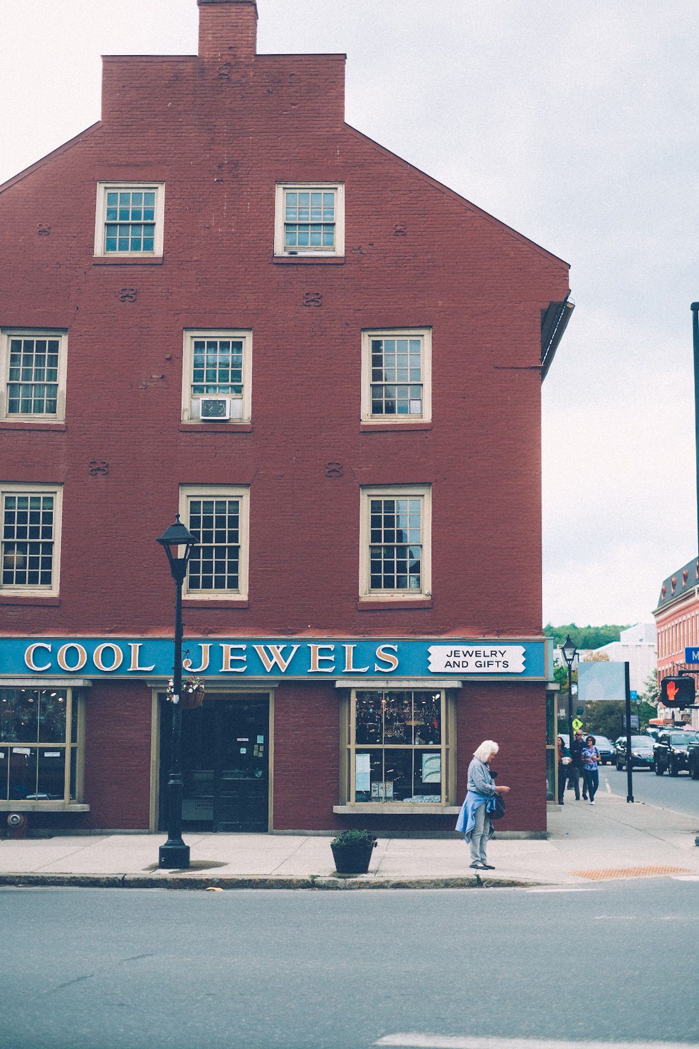 A woman stands on the street corner in front of a big burnt red building with a hand drawn sign that says Cool Jewels.