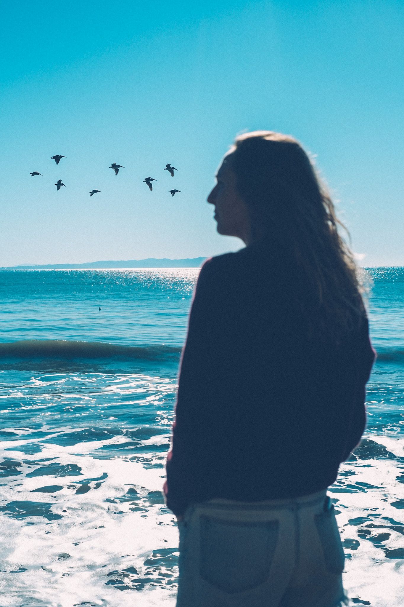 A woman with long flowing hair looks to the left of the photo as a flock of birds align with her eyesight, over the blue sea.