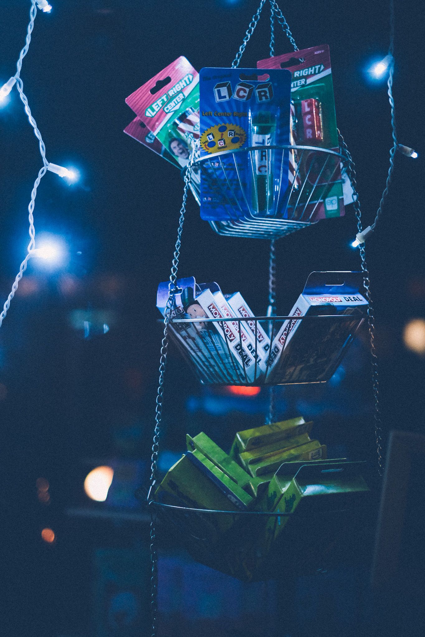 From inside the window of a game store at night, decks of card games hang in a three-tiered basket. Everything is bathed in blue light emanating from the strand of lights lining the windowsill.