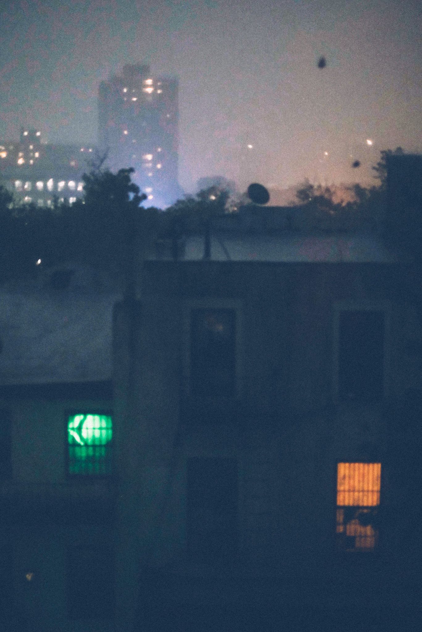 The view from inside an apartment, looking through a window out at a foggy, rainy night, buildings lit in the misty background with two windows glowing in the foreground, green and orange.