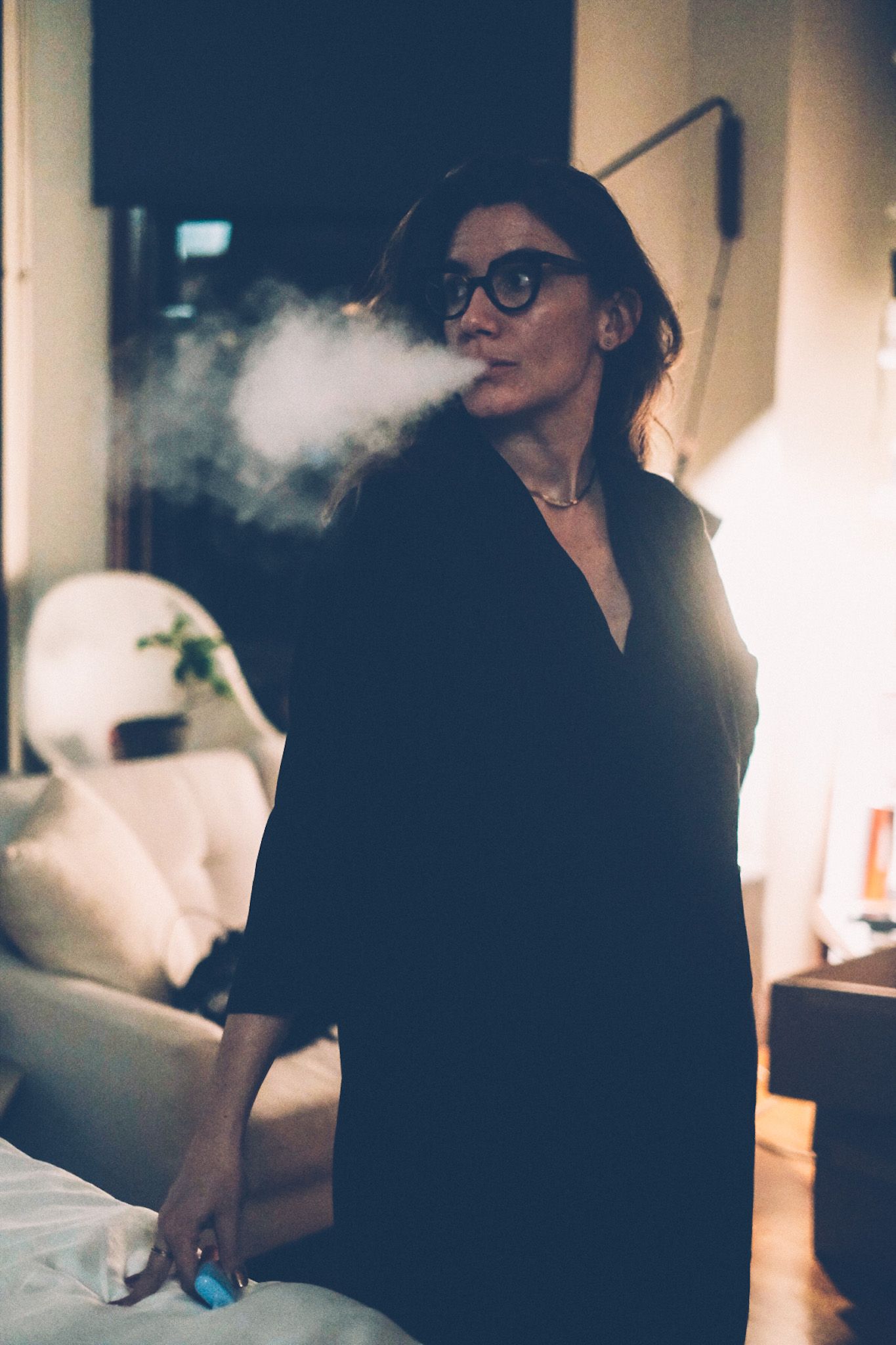 A woman stands in her apartment, looking over her shoulder as she blows smoke out of her mouth.