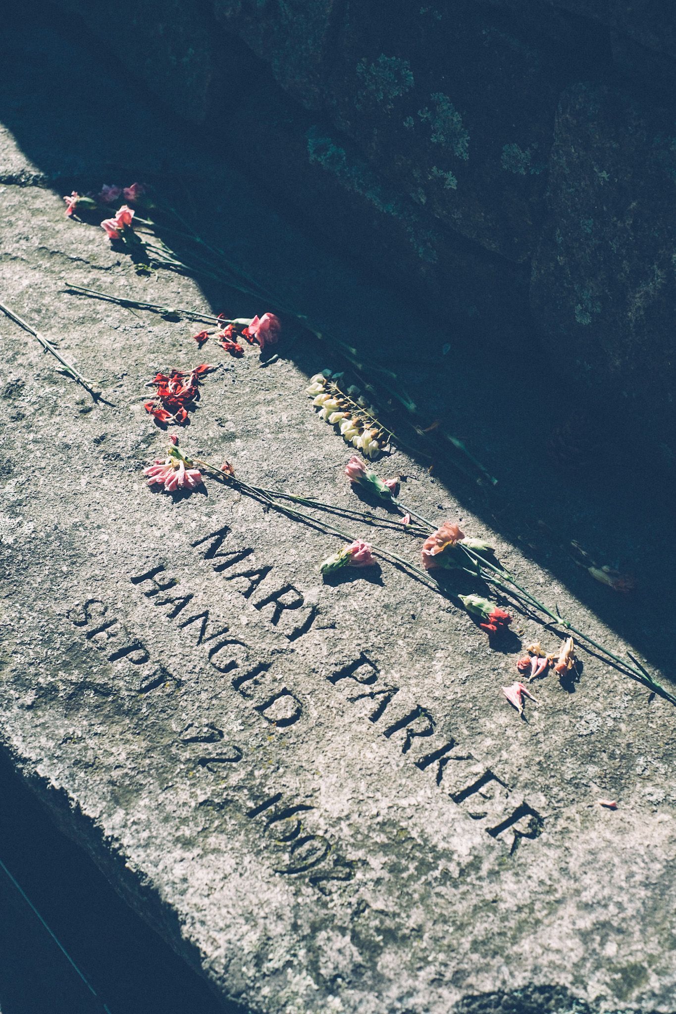 A gravestone in the harsh sunlight reads “Mary Parker / Hanged” with flowers scattered around it.