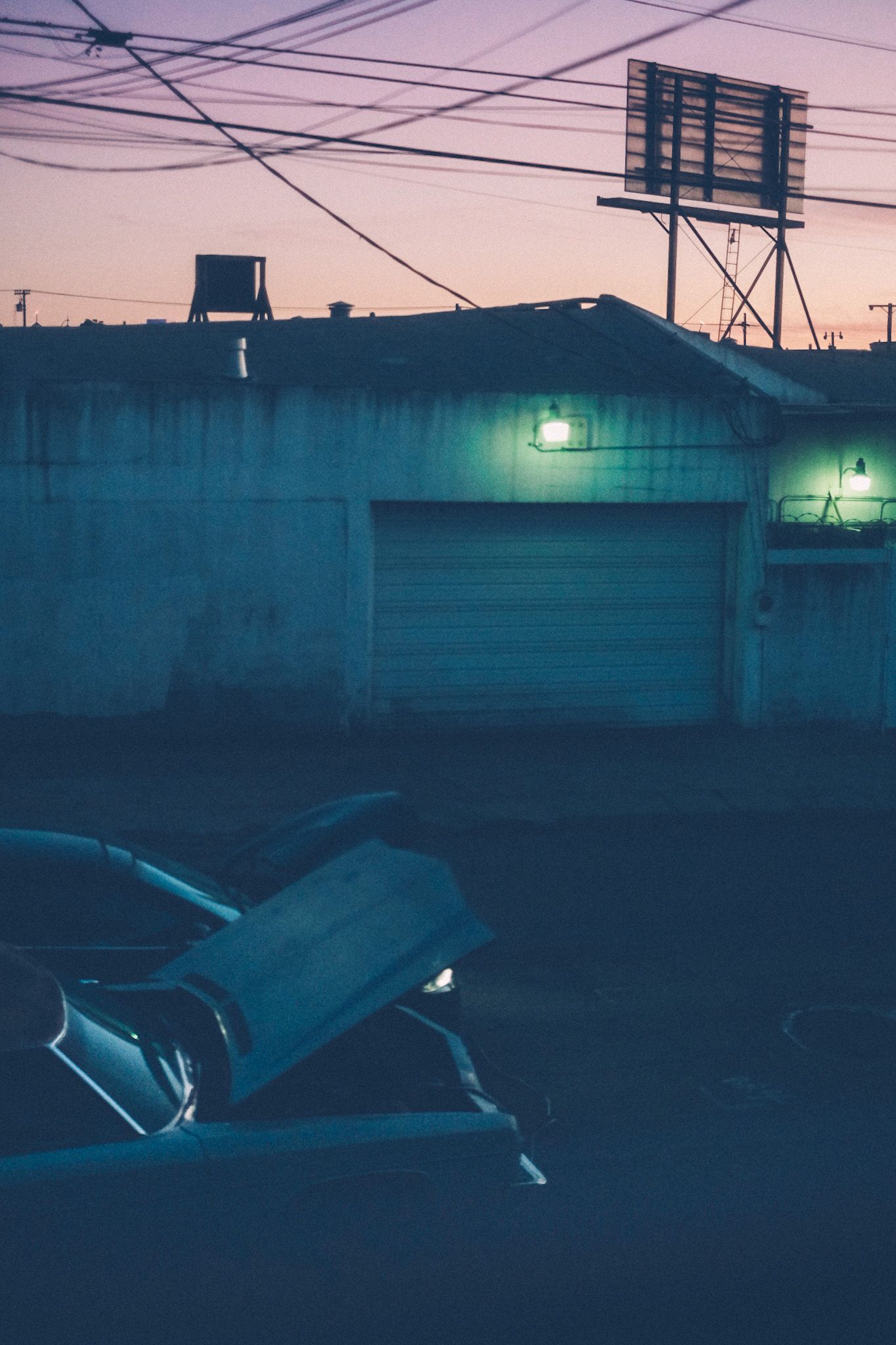 The hood of an old car is propped up as it gets jumped by another car, a green light of a garage across the street glowing against the pink sunset in the background. A billboard rises in the near distance, but you can only see its backside.