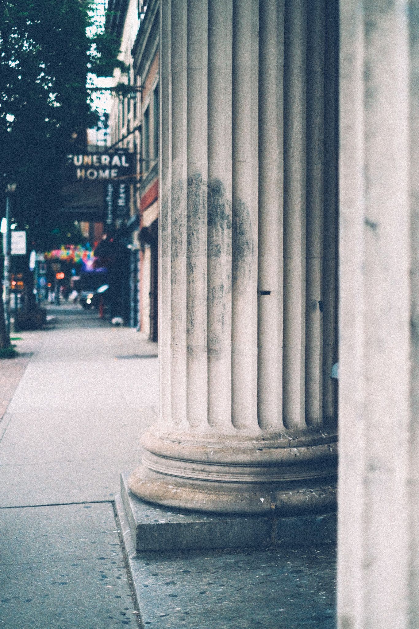 Dirty white pillars on a city street take up the right half of the frame, while in the distance the words “funeral home” line a sign.