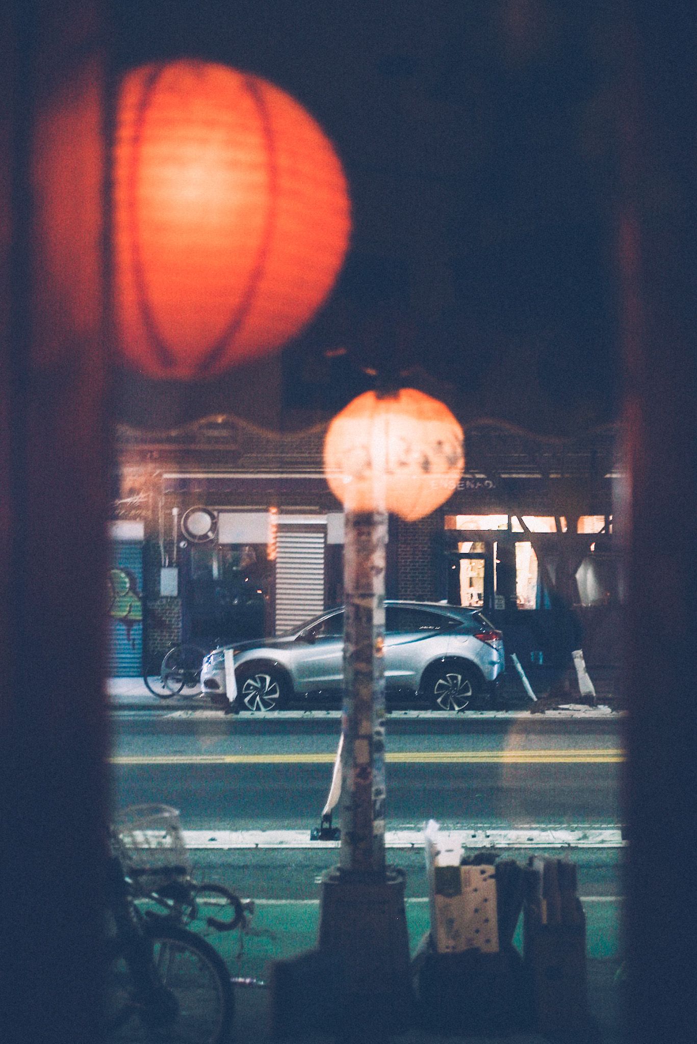 Through a window, the reflection of red paper lanterns is overlaid onto a street scene, bikes and boxes and sticker-covered phone piles and a car and buildings in the background. It is night.