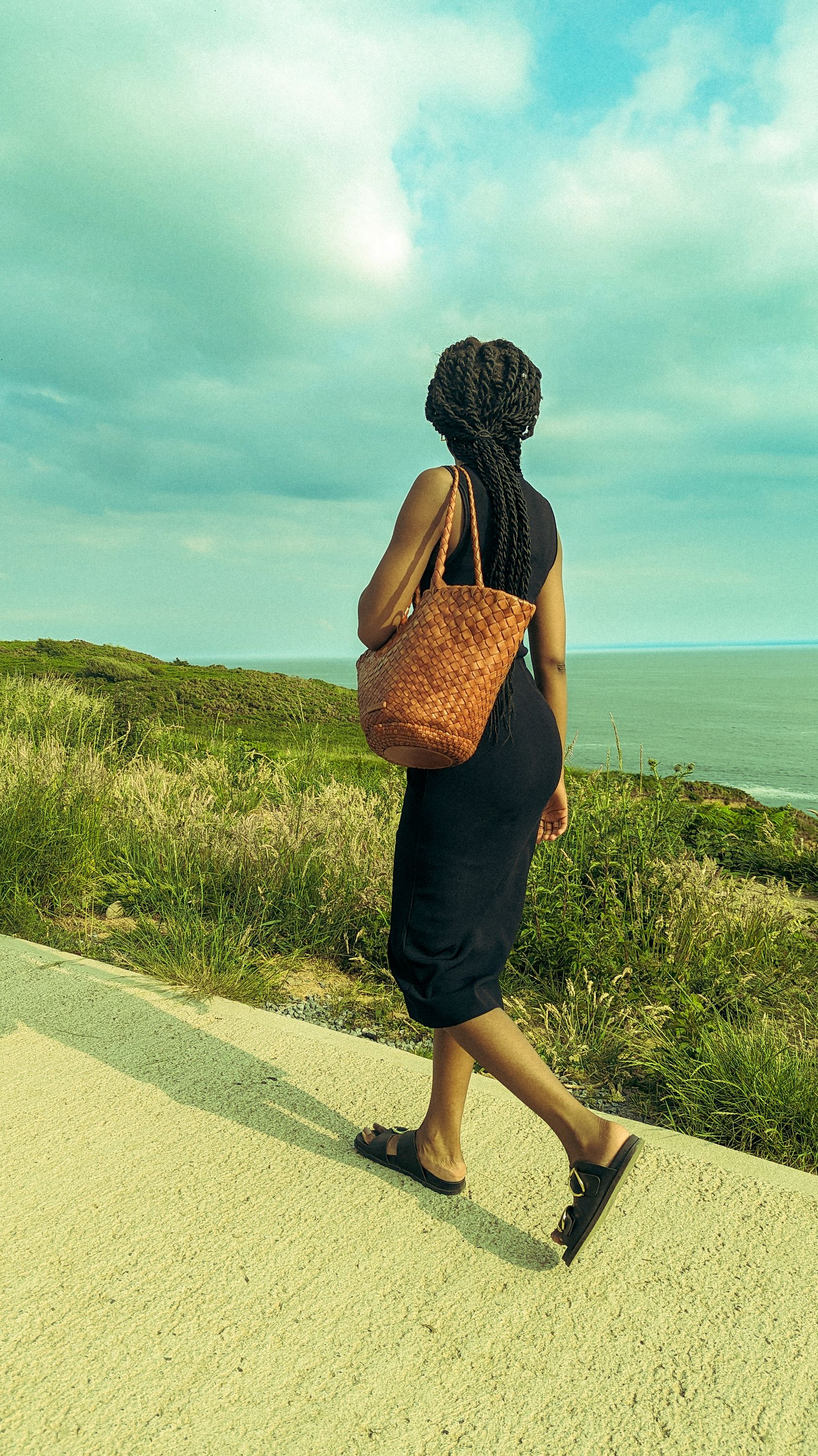 Back view of a woman with long hair, wearing a sleeveless dress and carrying a woven bag