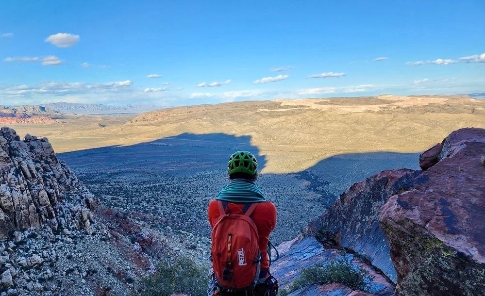 A person in a green helmet and orange backpack sits observing a vast desert landscape with mountains in the distance under a cloudy sky.
