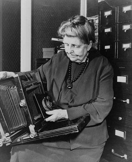 Frances Benjamin Johnston, three-quarter length portrait, holding and looking down at camera, facing slightly left