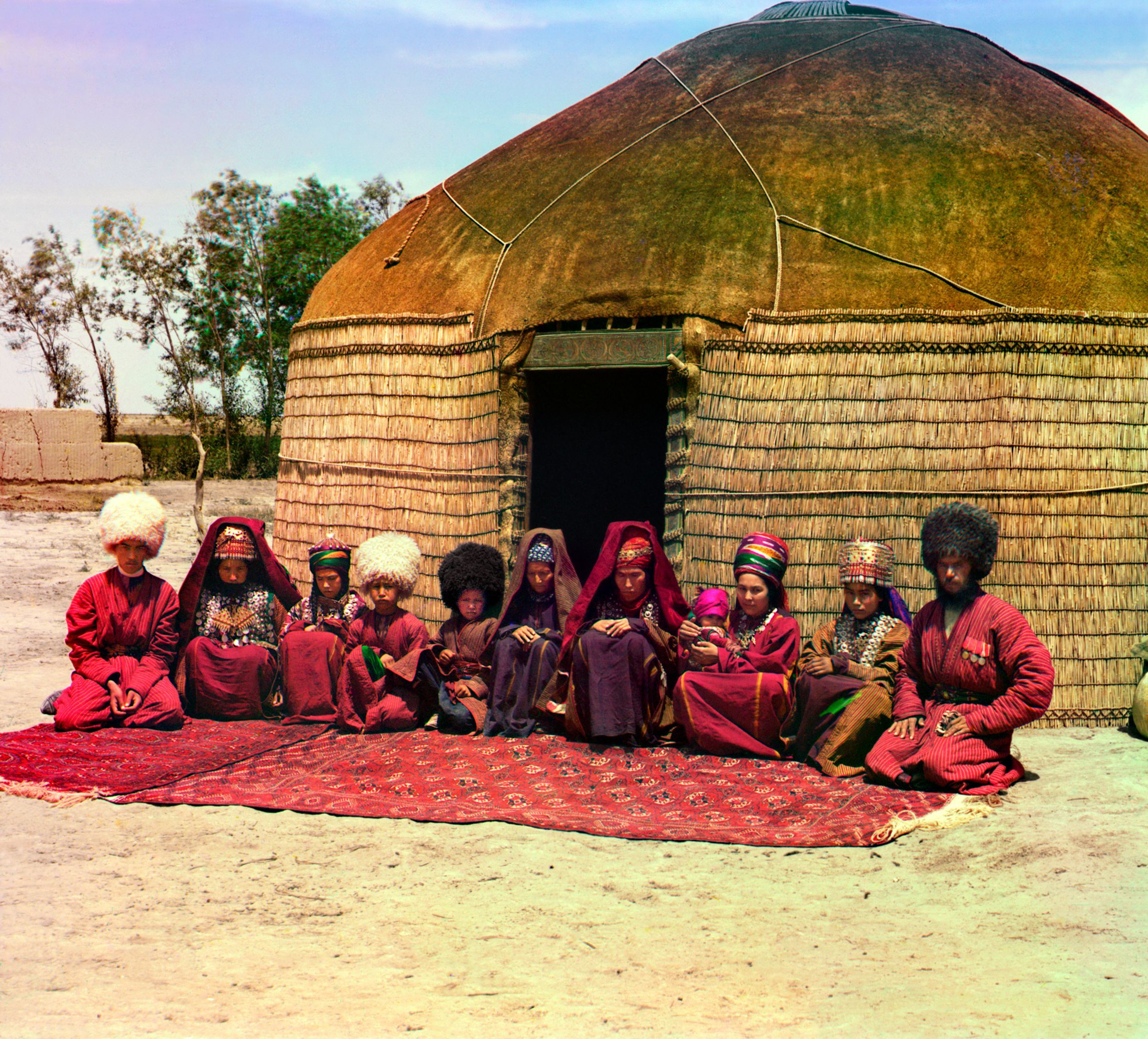 Group of eleven adults and children, seated on a rug, in front of a yurt