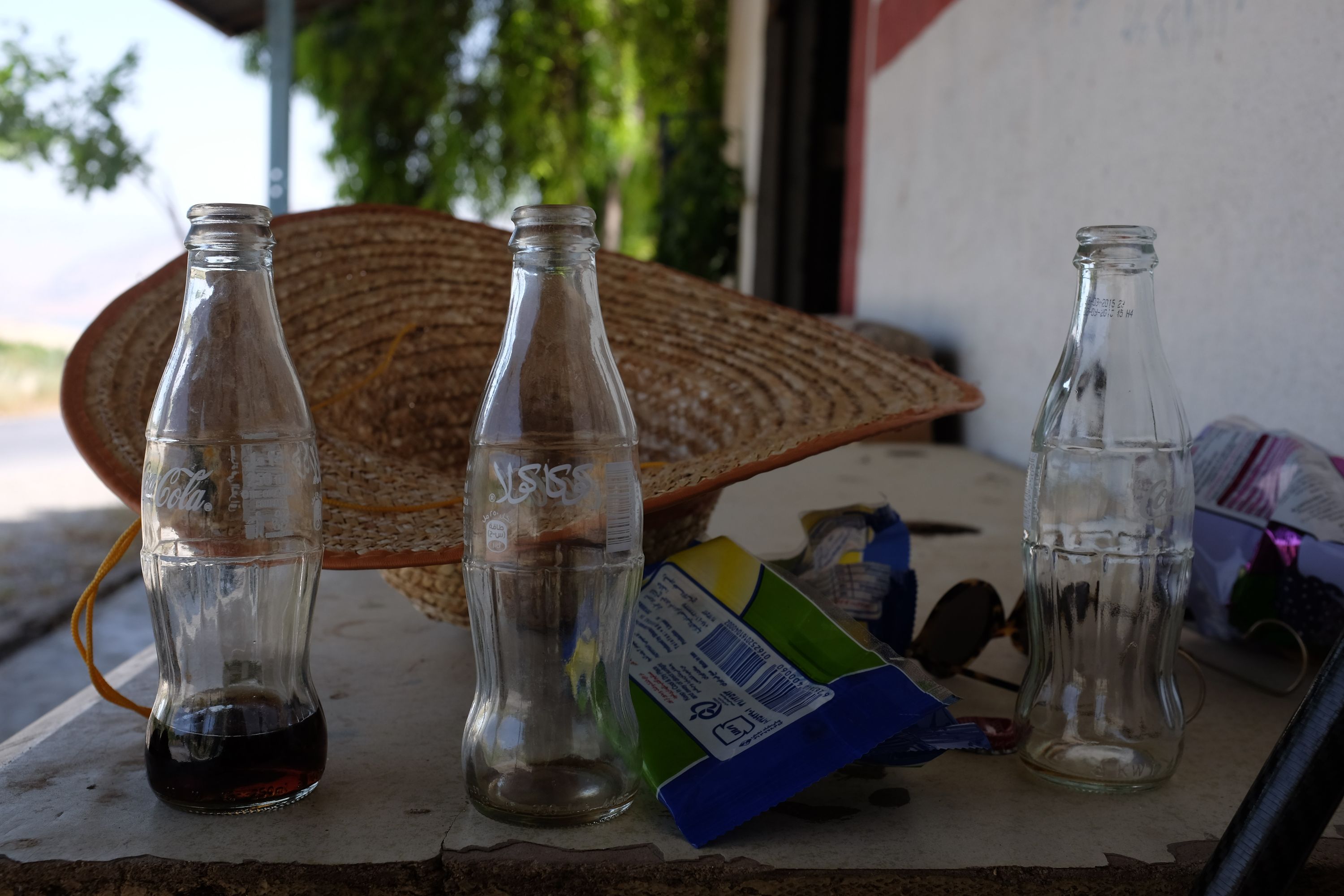 Three bottles of Coke, a little left in one of them, a large straw hat, and a pair of sunglasses on a table.