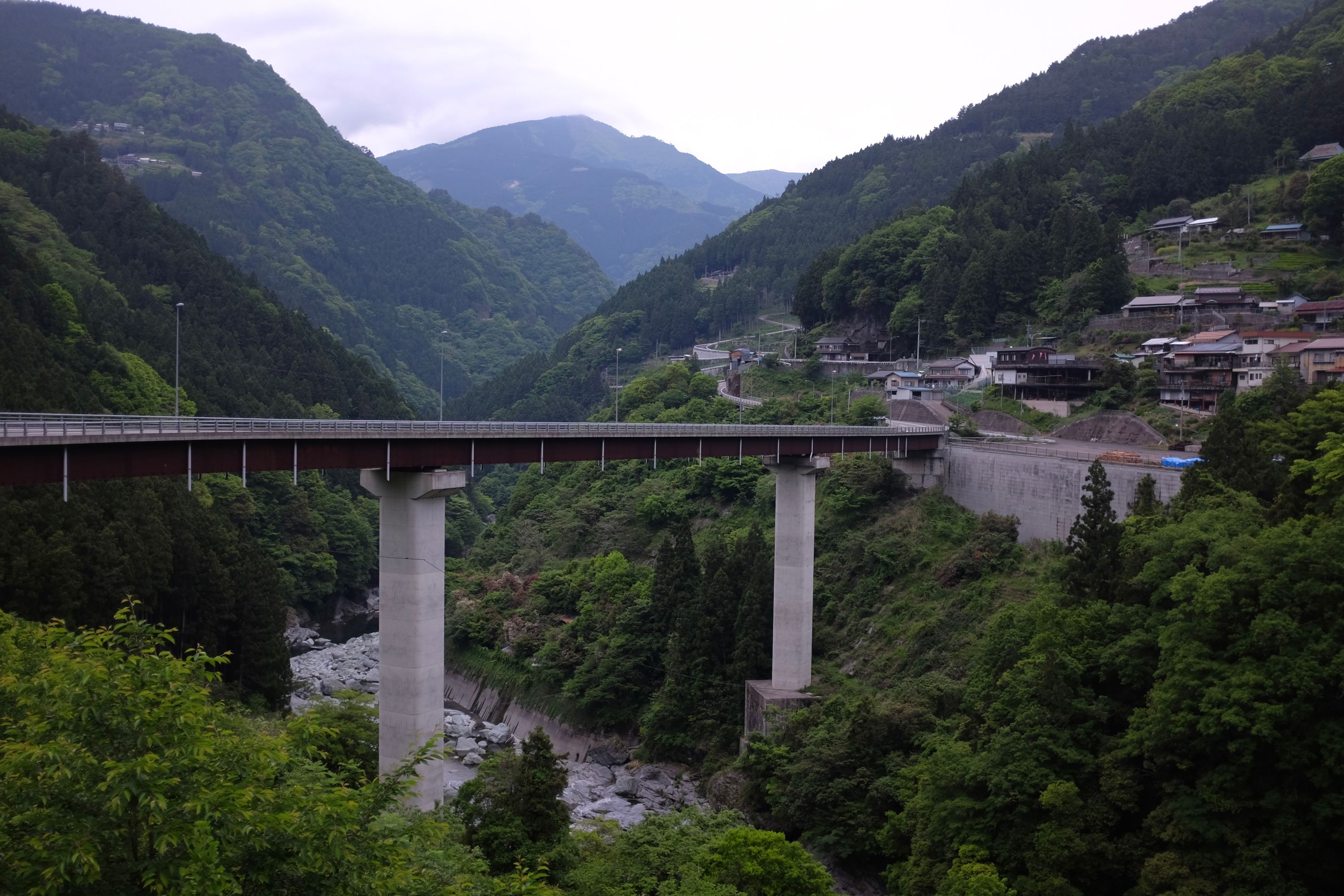 A highway crosses a mountain river on a high concrete bridge, with a village on the other side and forested hills in the distance.