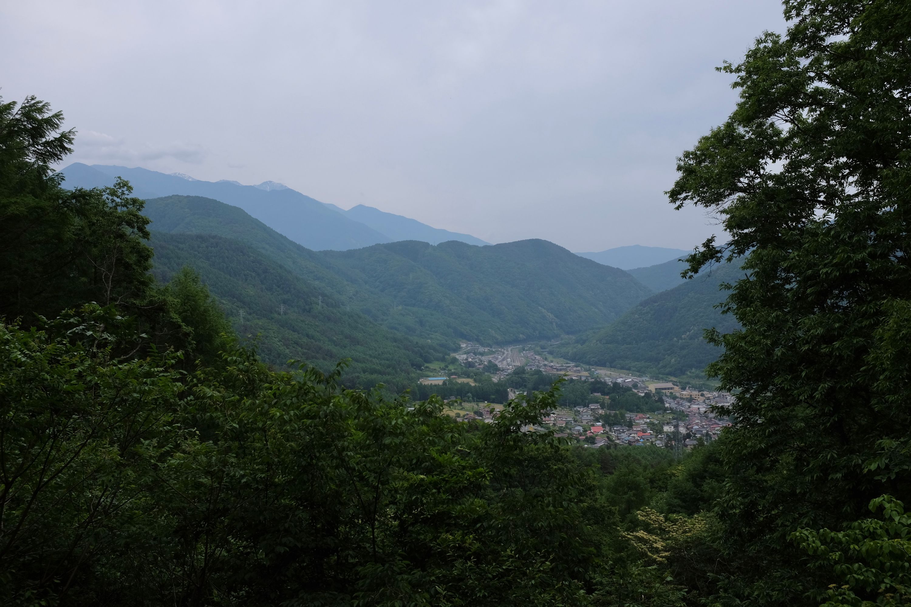 A view from a mountain pass reveals a village in a valley and snow-covered mountains in the distance.