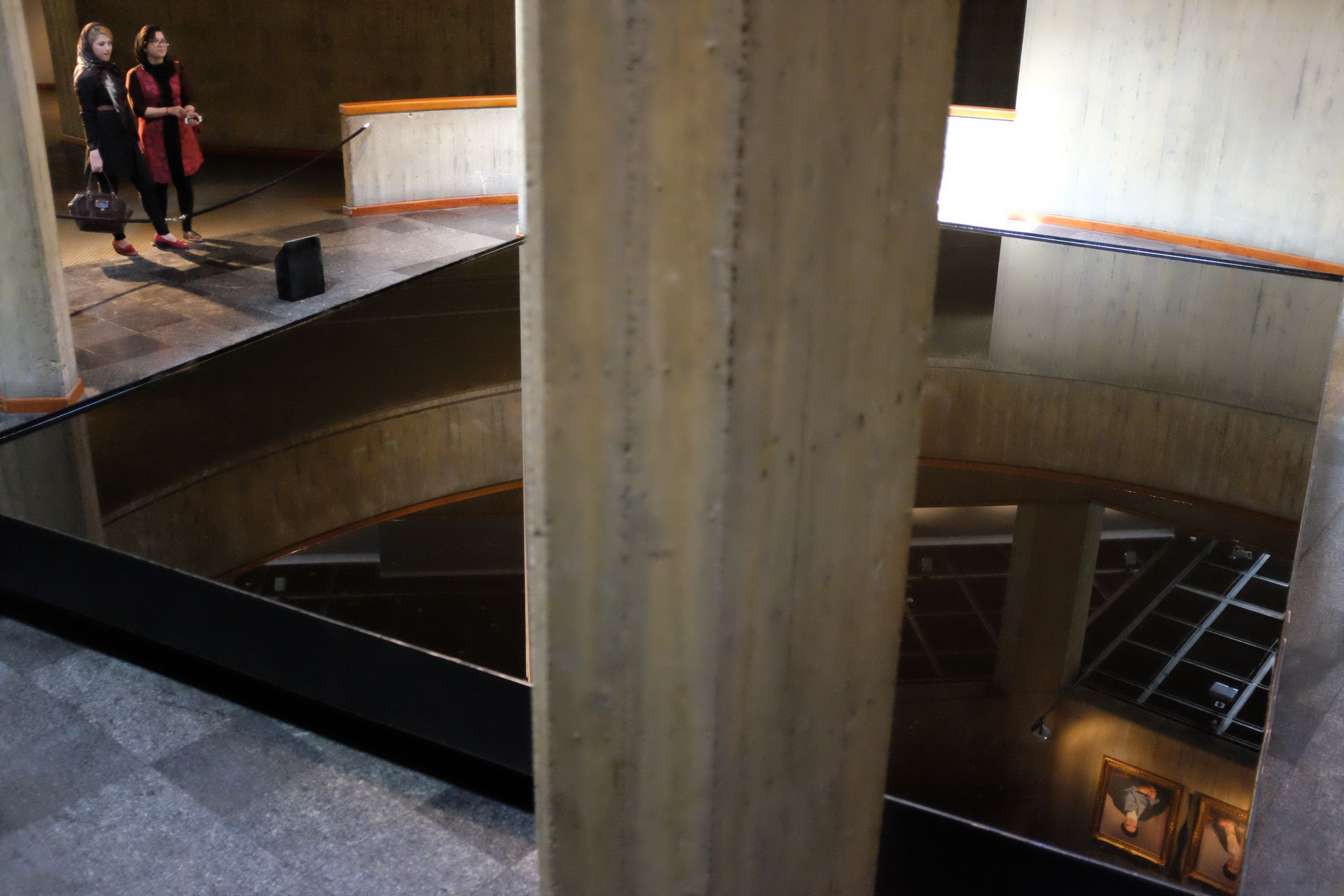 Two women look into a large pool of crude oil, a 1970s piece of modern art, in which portraits of the two Supreme Leaders of Iran are reflected.