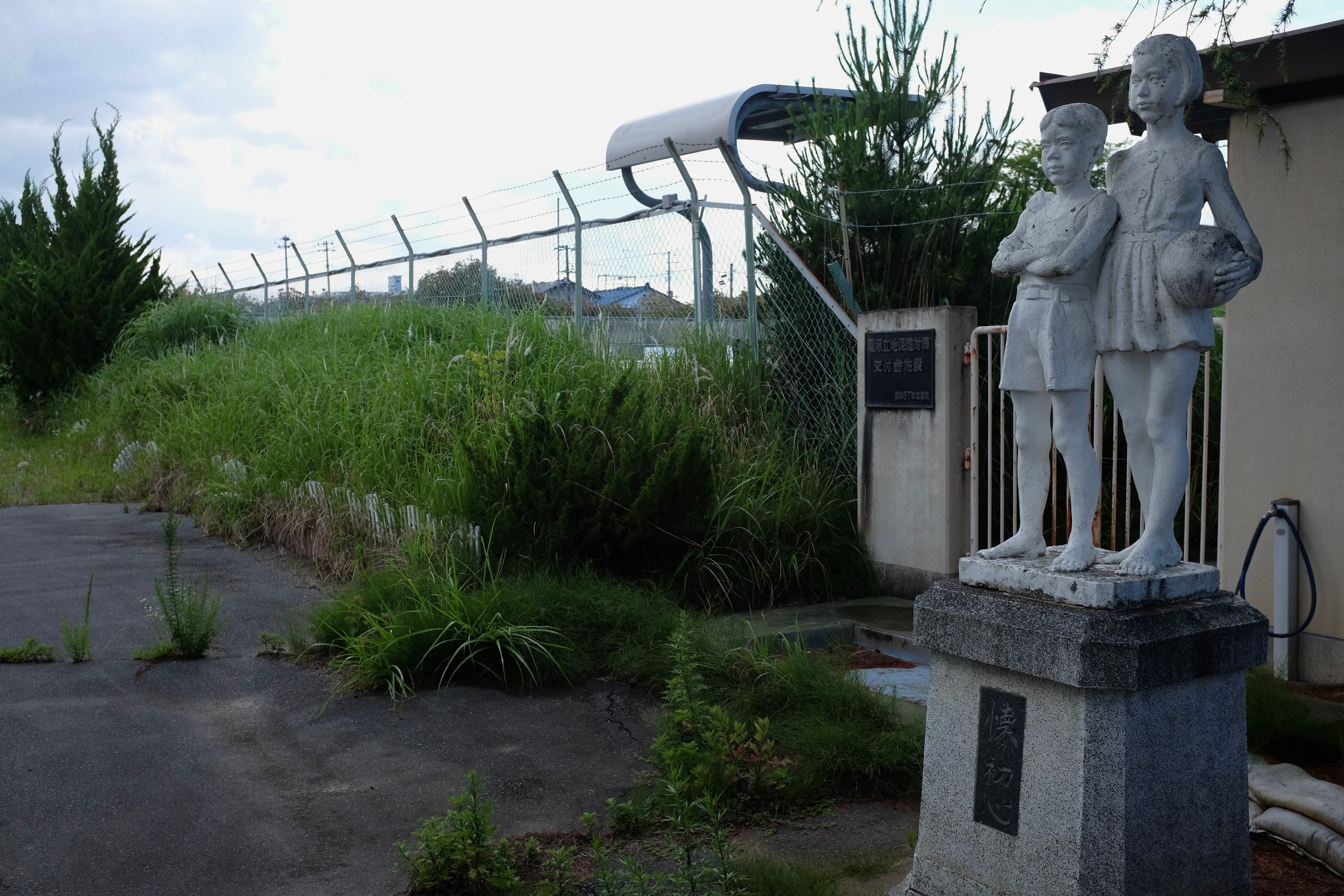 A statue of a schoolgirl and a younger schoolboy in an school yard overgrown with weeds.