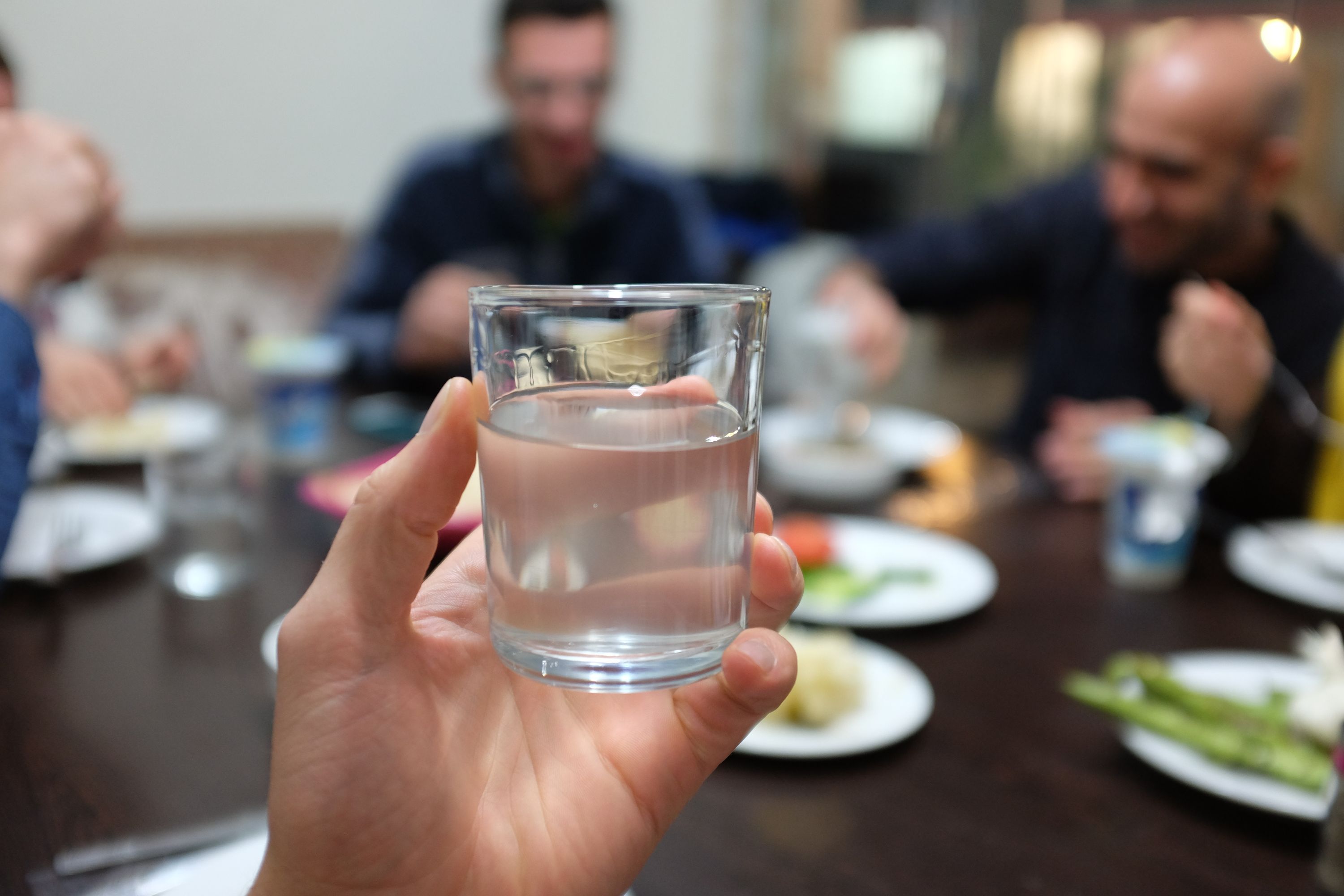 A hand hold a glass of clear liquid whose viscosity suggests it is hard liquor. Behind the glass, people sit around a dinner table.