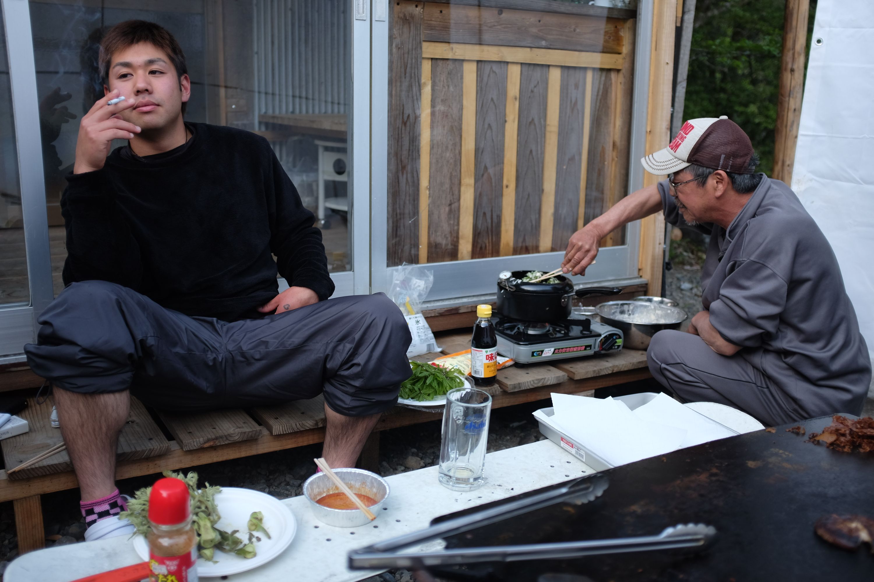 Two men by a cabin, one of them relaxing with a cigarette, the other frying vegetables on a gas burner.