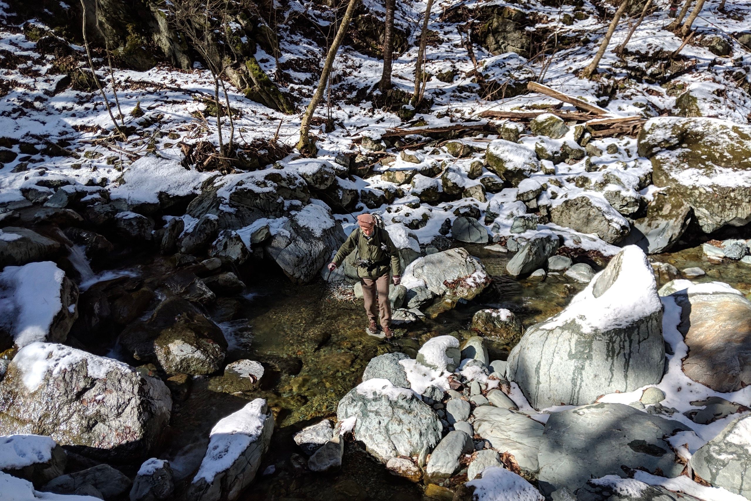 Looking down from above, a man, the author, makes its way across a stream, balancing on rocks.