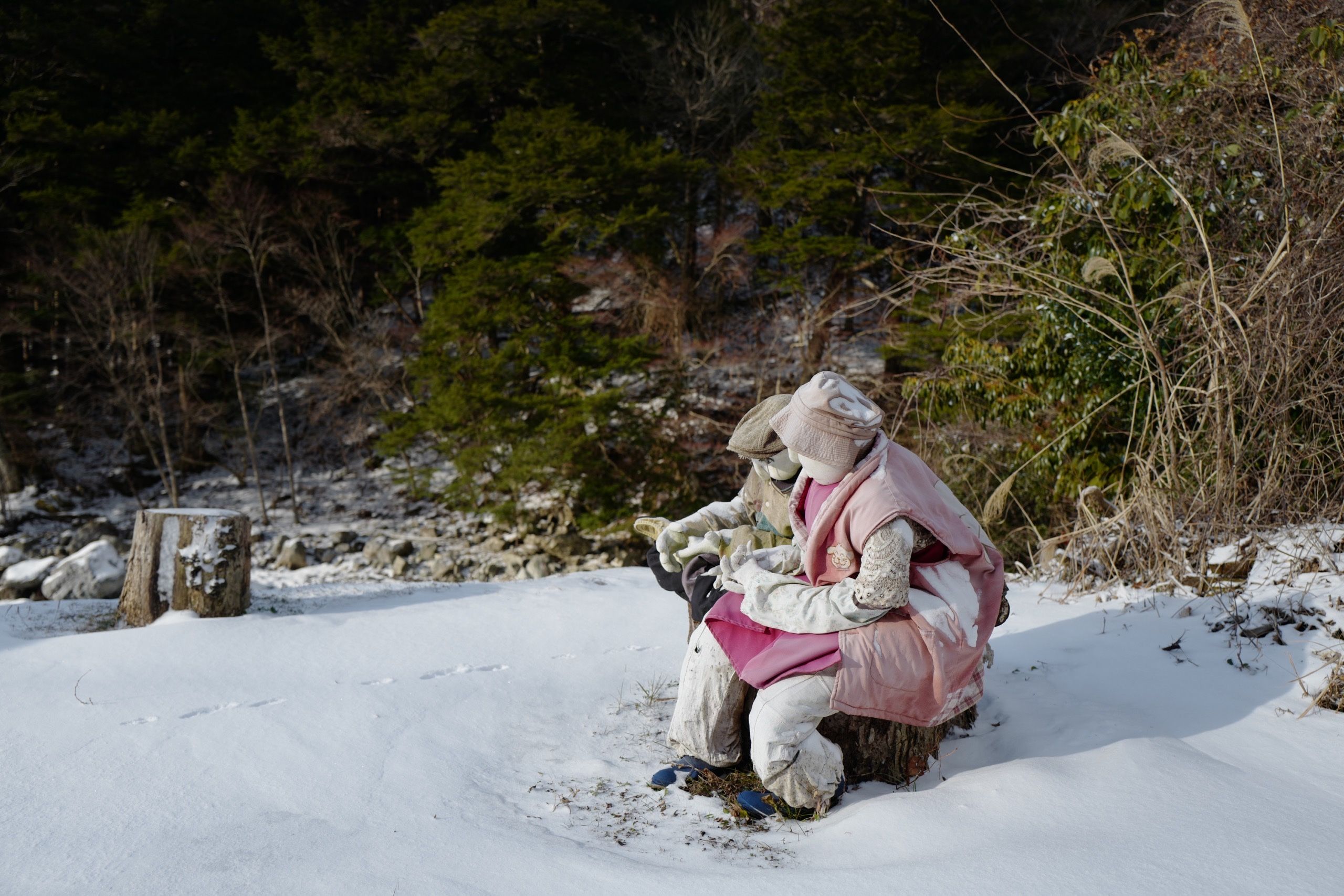Another two dolls sit outside on a bench in the snow.