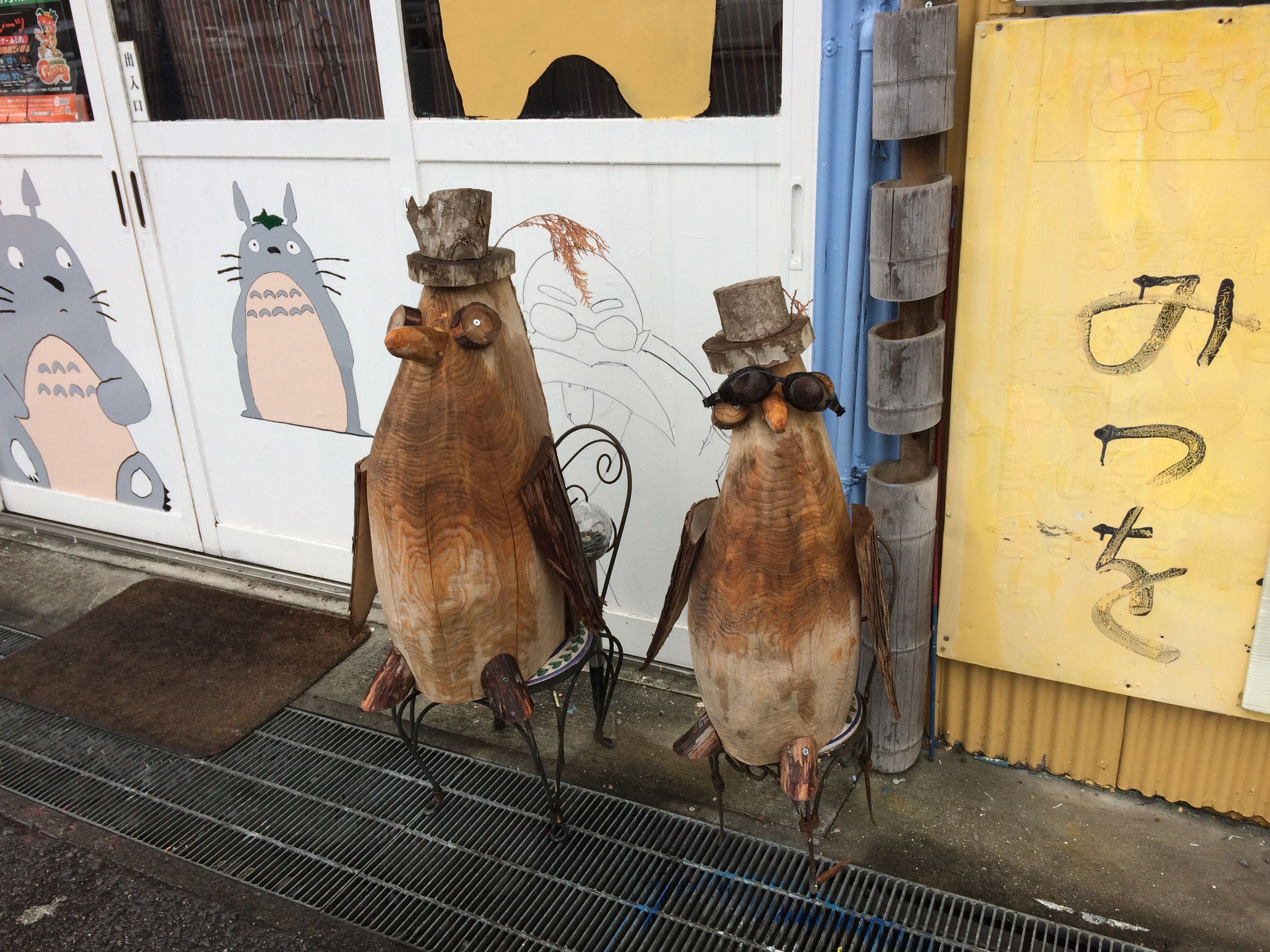 Two carved wooden birds in top hats, the female in sunglasses, sit on metal chairs outside a shop.