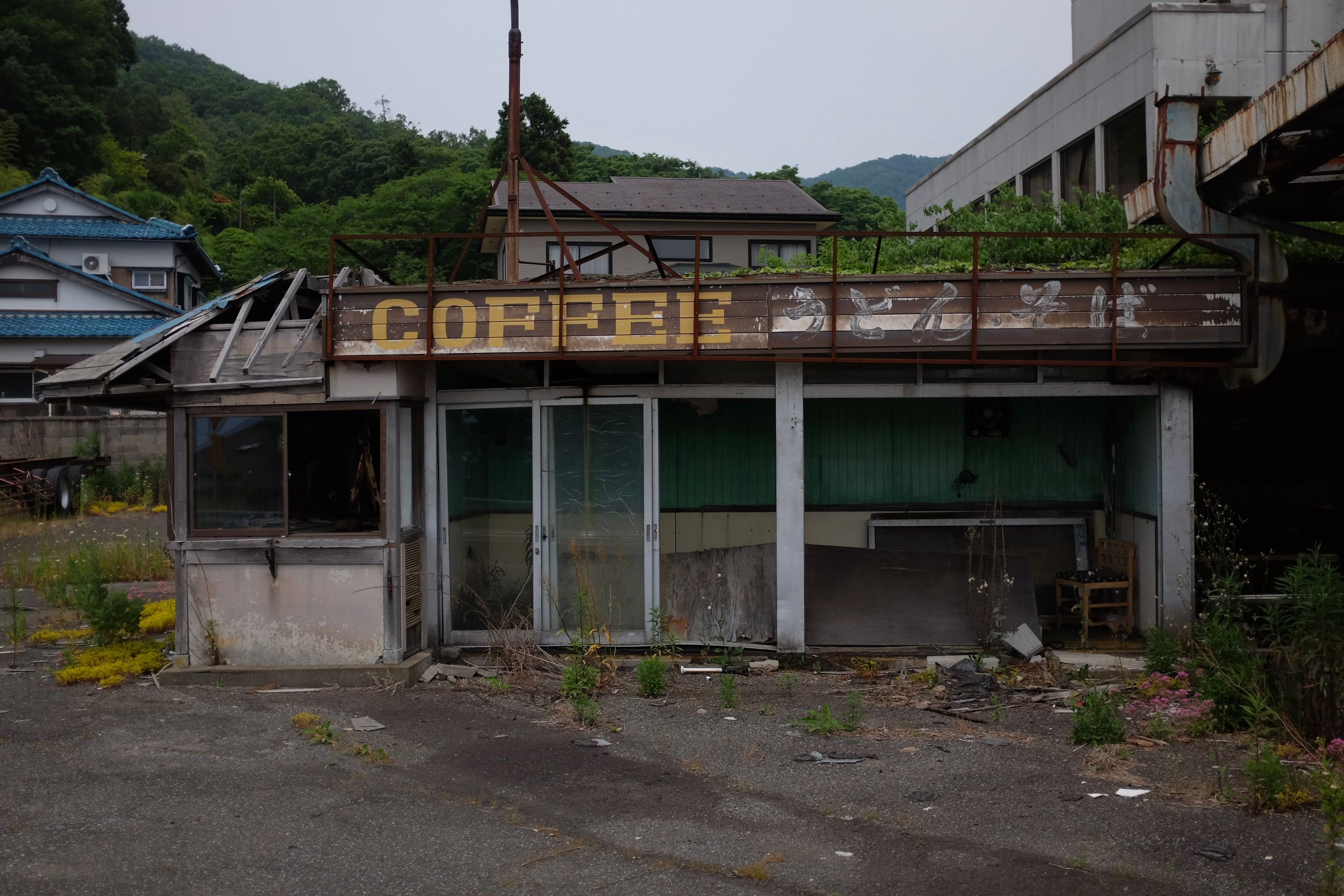 A dilapidated café with village houses and forested hills behind it.