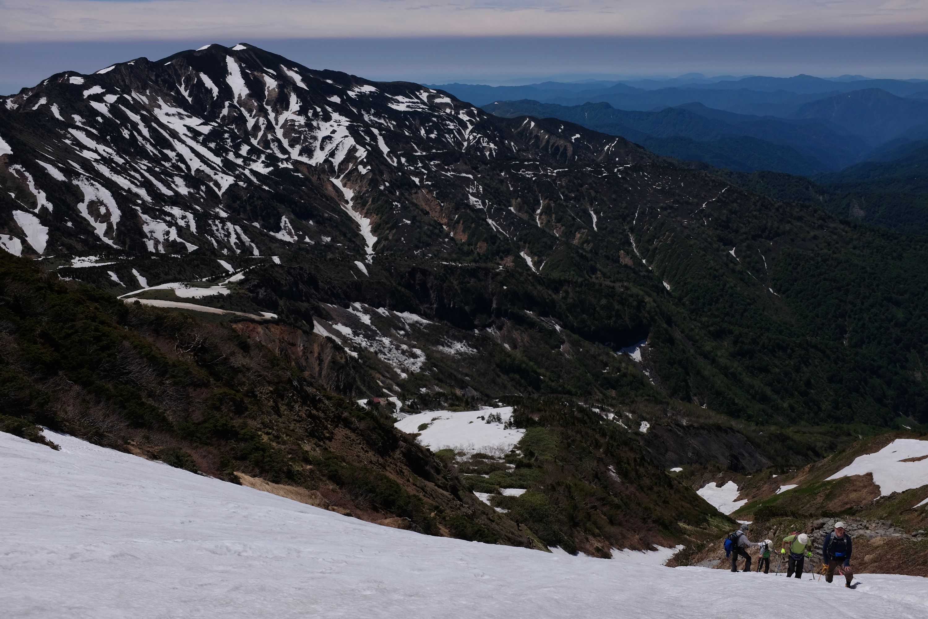 Climbers ascend a steep snowslope against a mountain scenery stretching to the horizon.