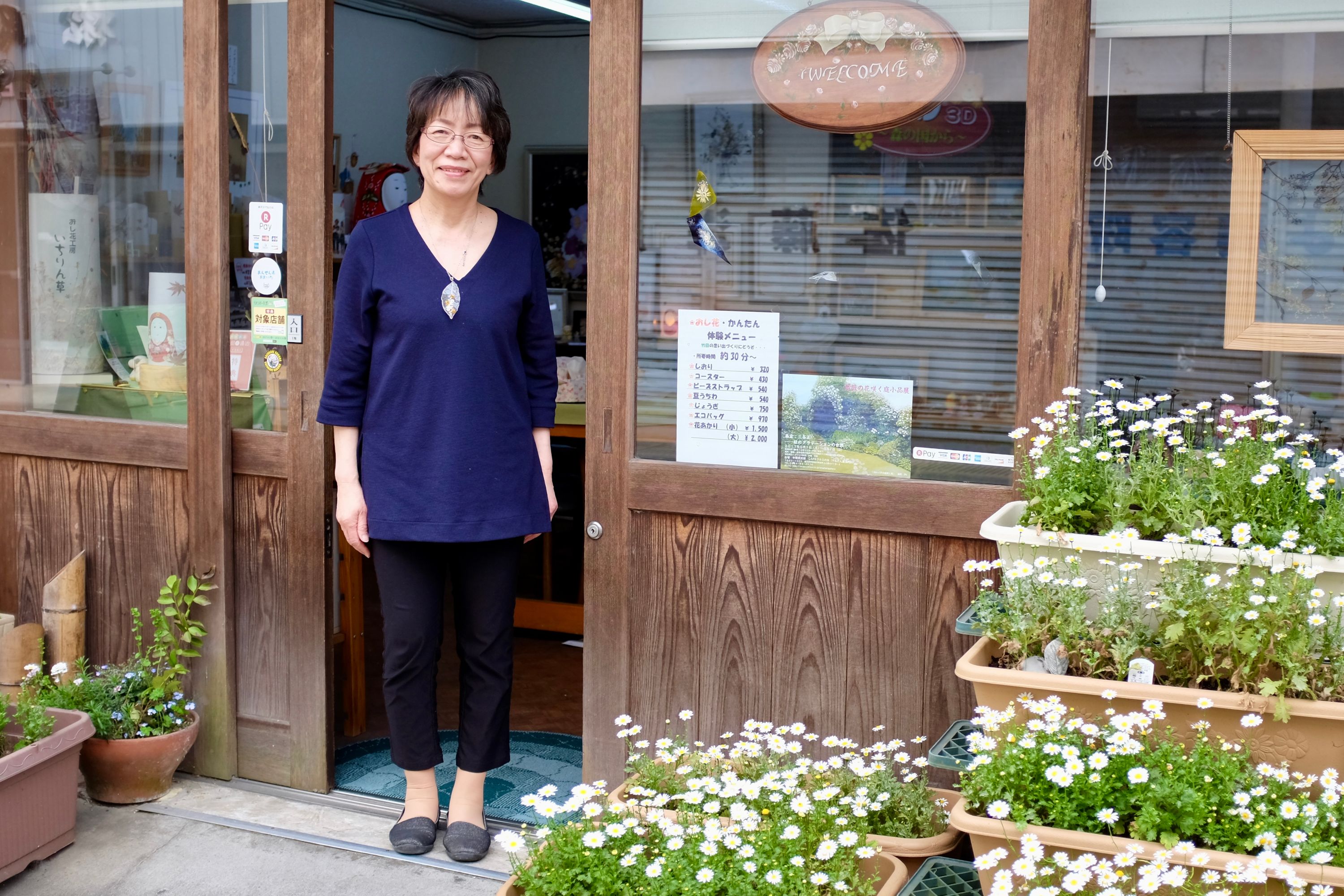 A middle-aged Japanese woman in glasses stands in the doorway of her shop, with many pots of live flowers outside.