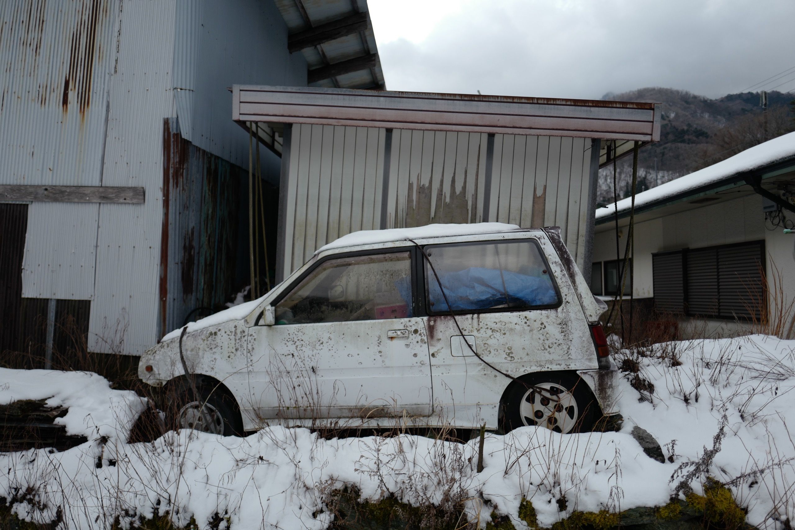 A small, dilapidated white cars parked in a snowy back yard between shacks.