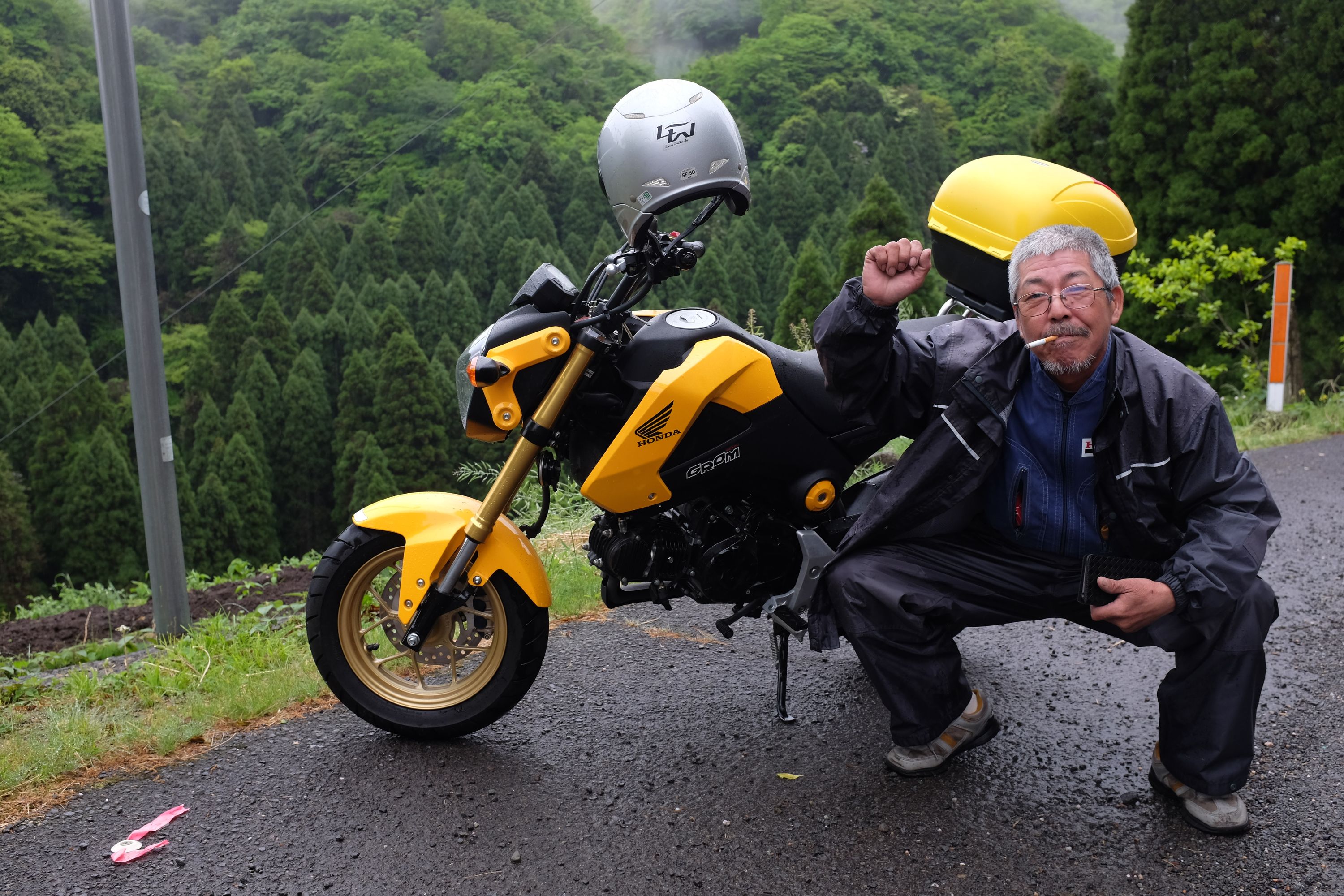 The same man squats by his motorcycle, raising a fist and looking very happy, his cigarette sticking out of his mouth.