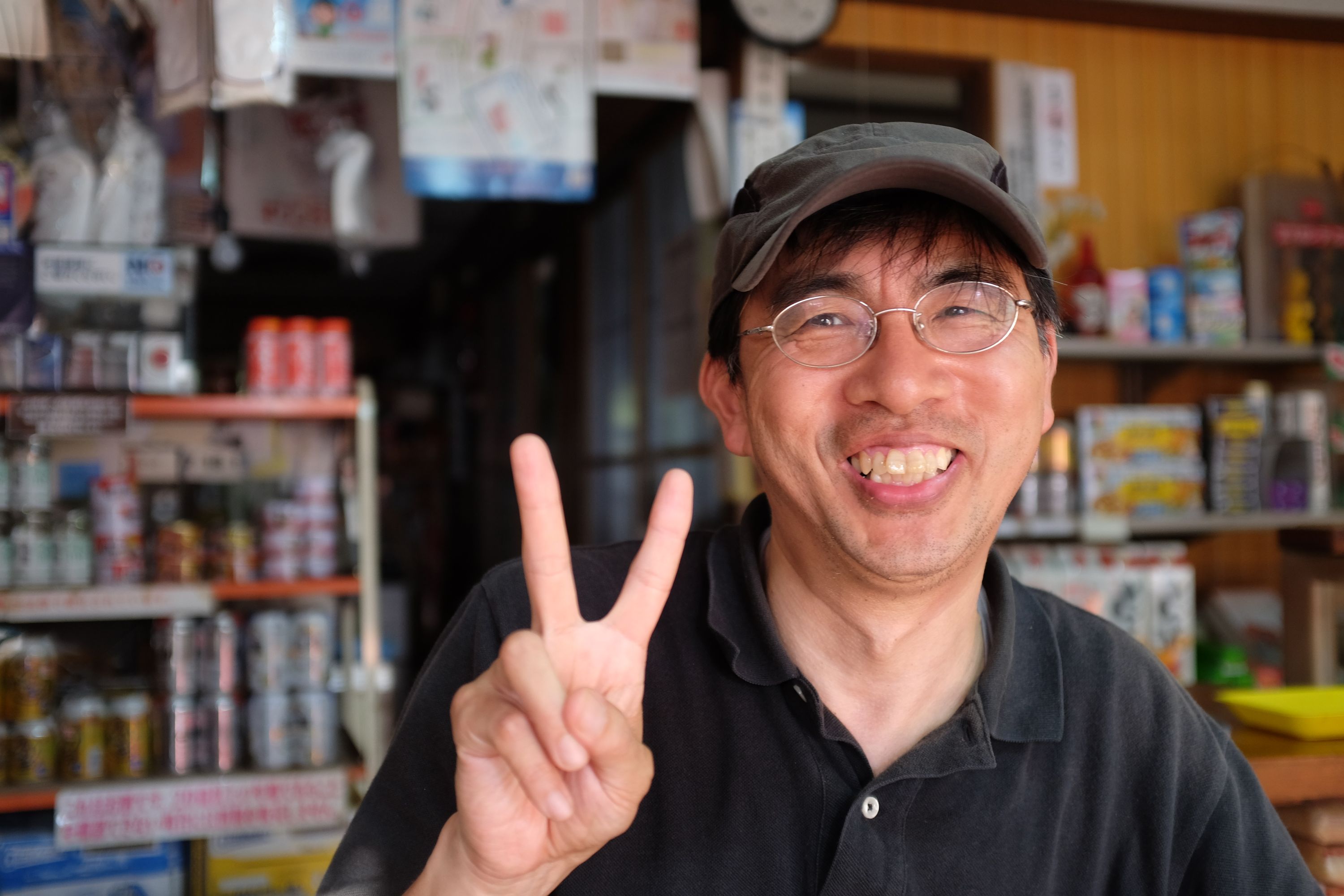 A Japanese man in glasses and a black polo shirt, Mr. Tadashi, smiles into the camera and shows the V sign.