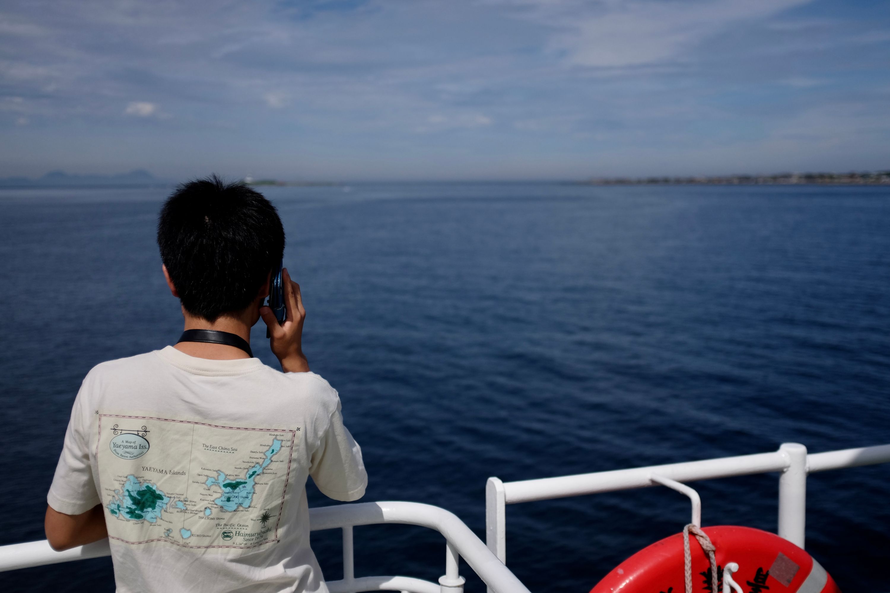 A man at the railing of a ferry looks out at the sea, wearing a t-shirt decorated with Ishigaki and Iriomote, two of Japan’s southernmost islands, thousands of kilometers away.