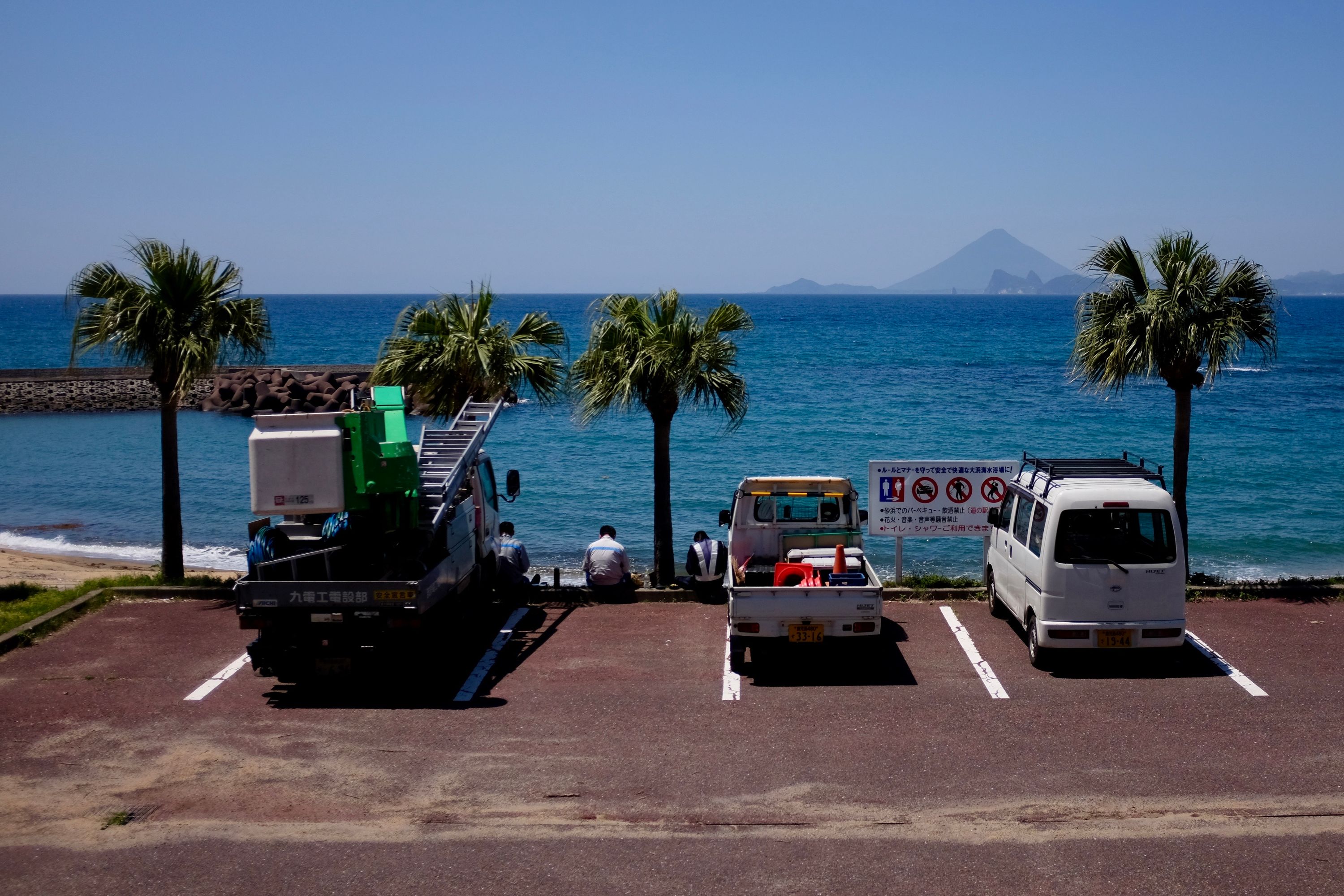 Three men eat lunch by their pickup trucks in a parking lot with an amazing view of Kagoshima Bay and Mount Kaimon.