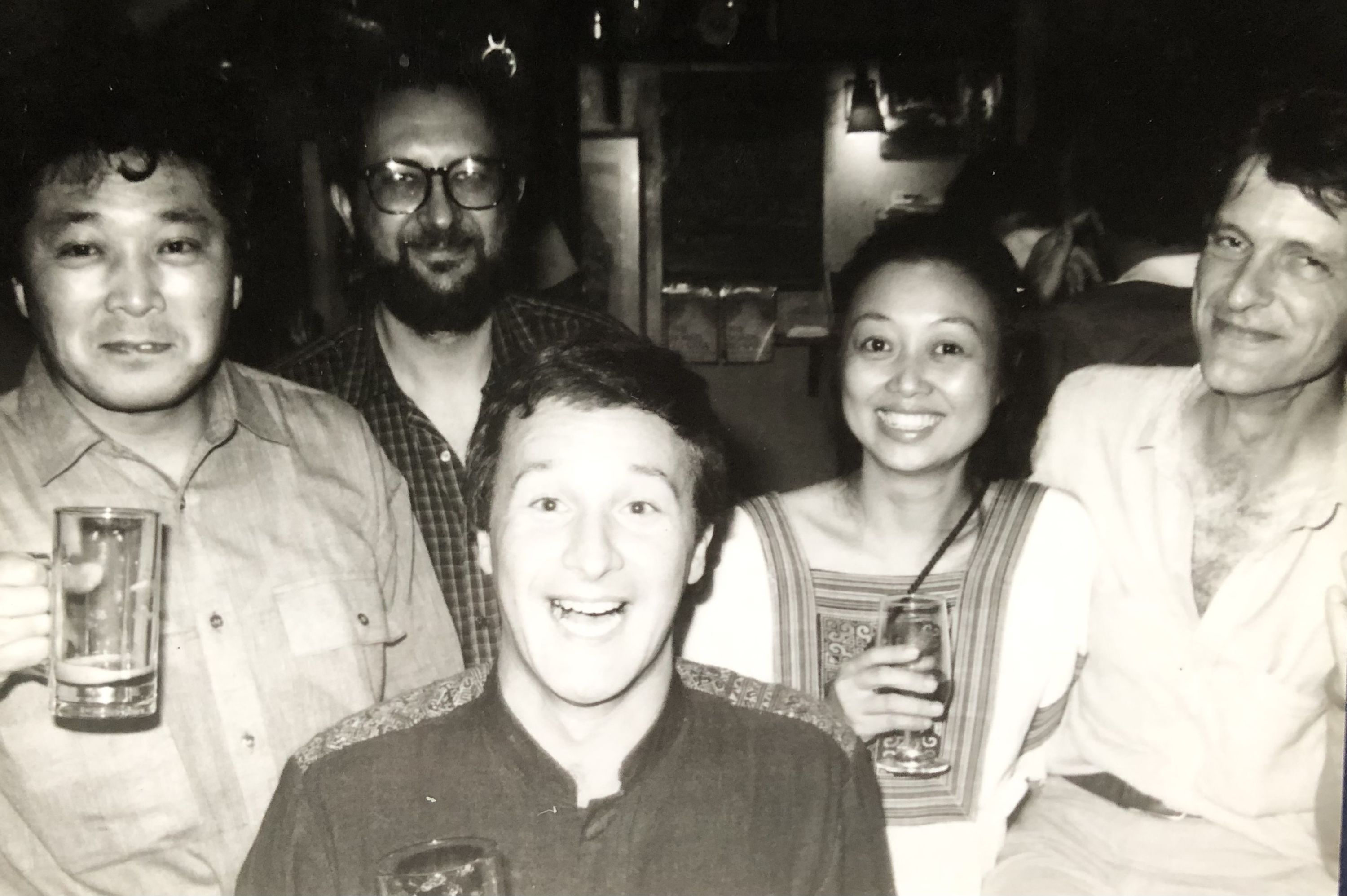 A black and white photograph showing five people gathered around a table in what appears to be a cozy bar or restaurant. From left to right: a young Asian man in a light-colored shirt holding a tall glass of beer, a bearded man wearing glasses and a plaid shirt, Alan Booth, an Englishman in the center foreground wearing a dark shirt with a cheerful expression, Su-chzeng Ong, Alan’s wife, a Malay-Chinese woman in a striped top holding a glass, and Timothy Harris, another Englishman on the right. The setting has warm lighting with bottles visible on shelves in the background, creating an intimate social atmosphere typical of 1980s Tokyo nightlife.