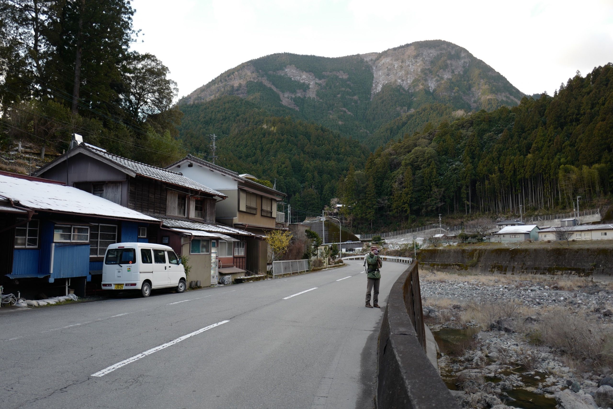 A man in winter clothes, the author, stands on a village road in front of a forested mountain.