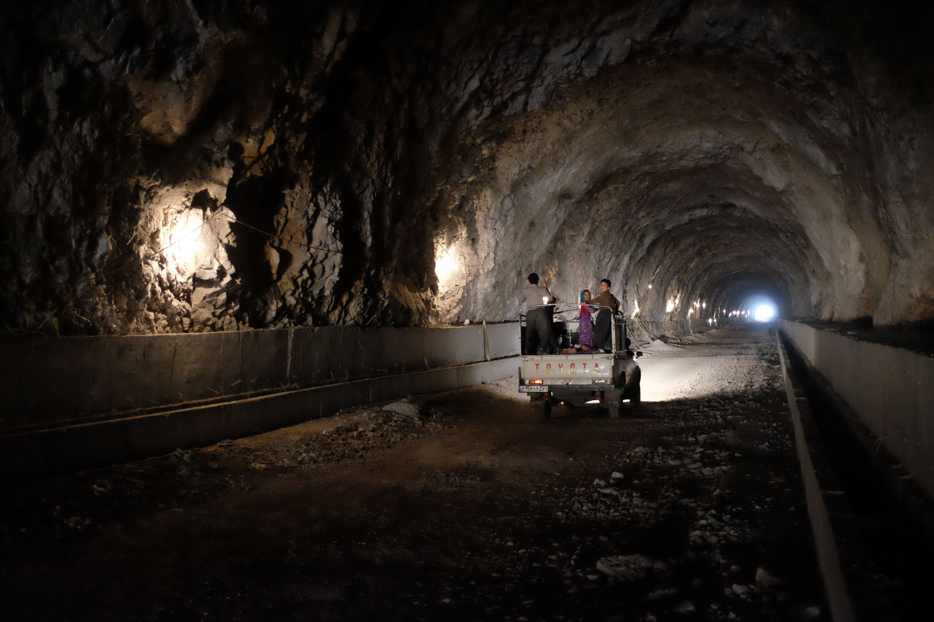 Children ride in the bed of a white Toyota pickup truck which drives along the dirt floor of long tunnel.