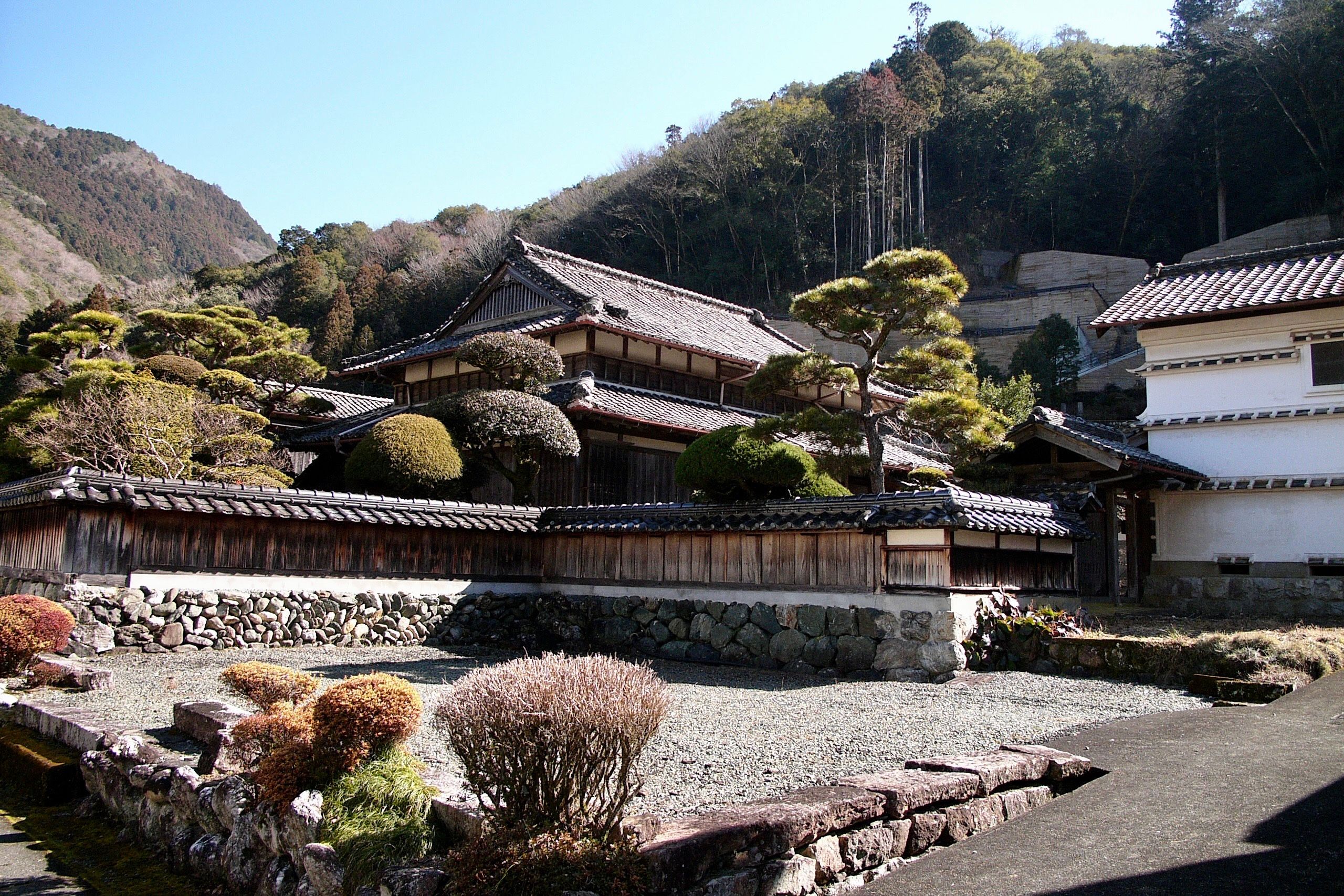 A traditional Japanese house in a village, complete with stone wall and carefully manicured trees.