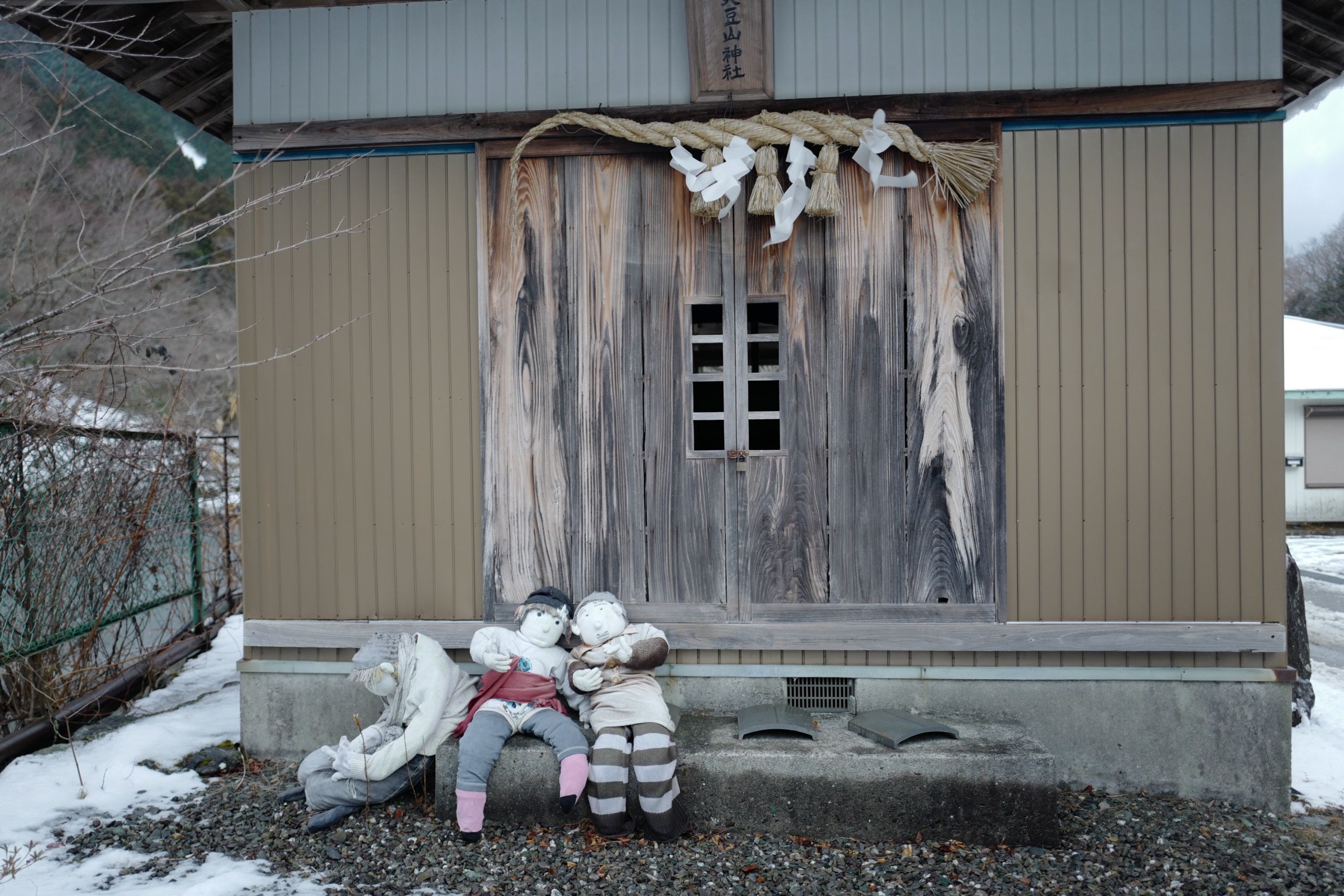 Three human-sized dolls sit on the porch of a Japanese shrine.