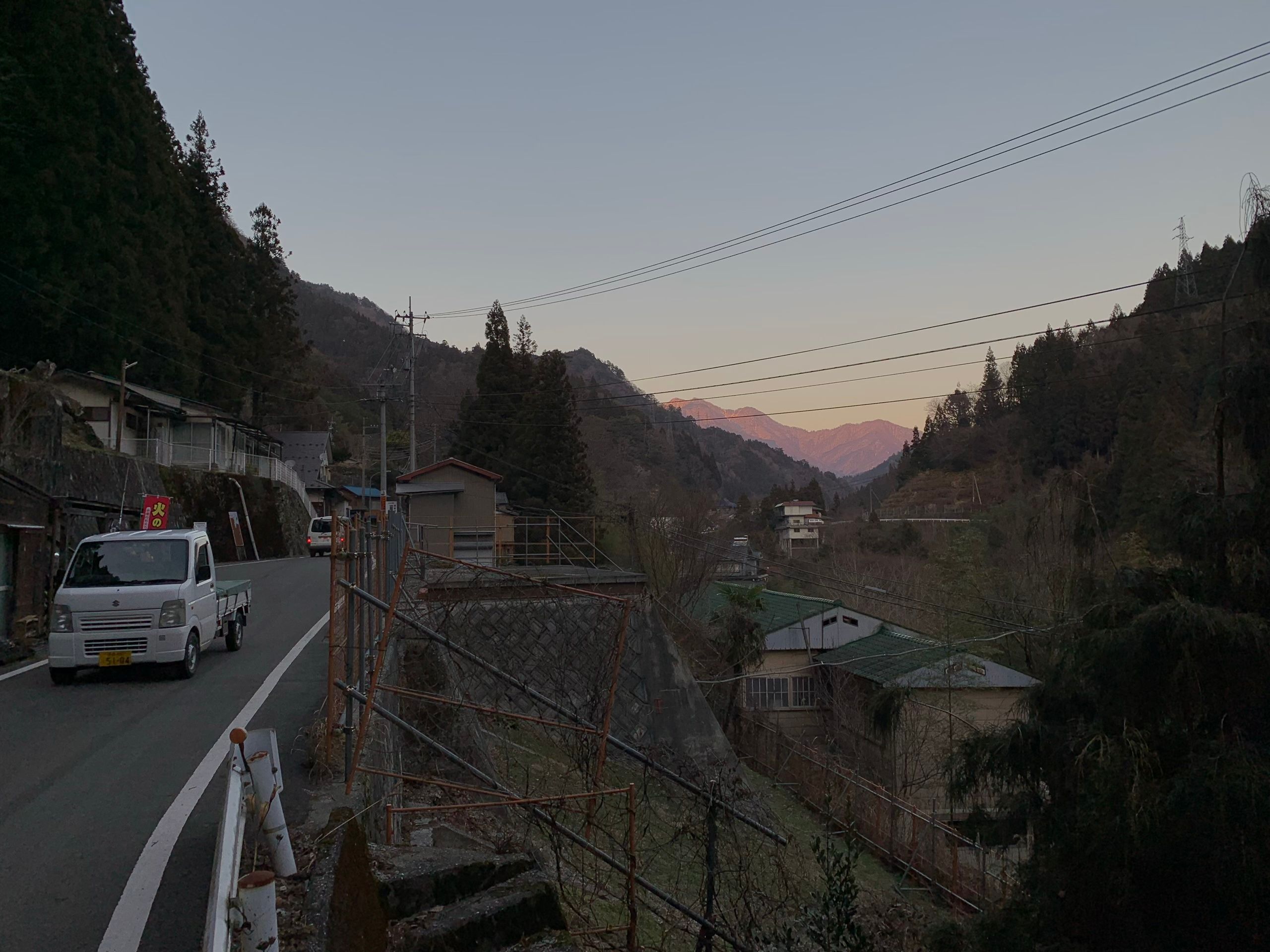 A very small van drives down a very narrow country road in a valley, with mountains on the horizon lit with alpenglow.